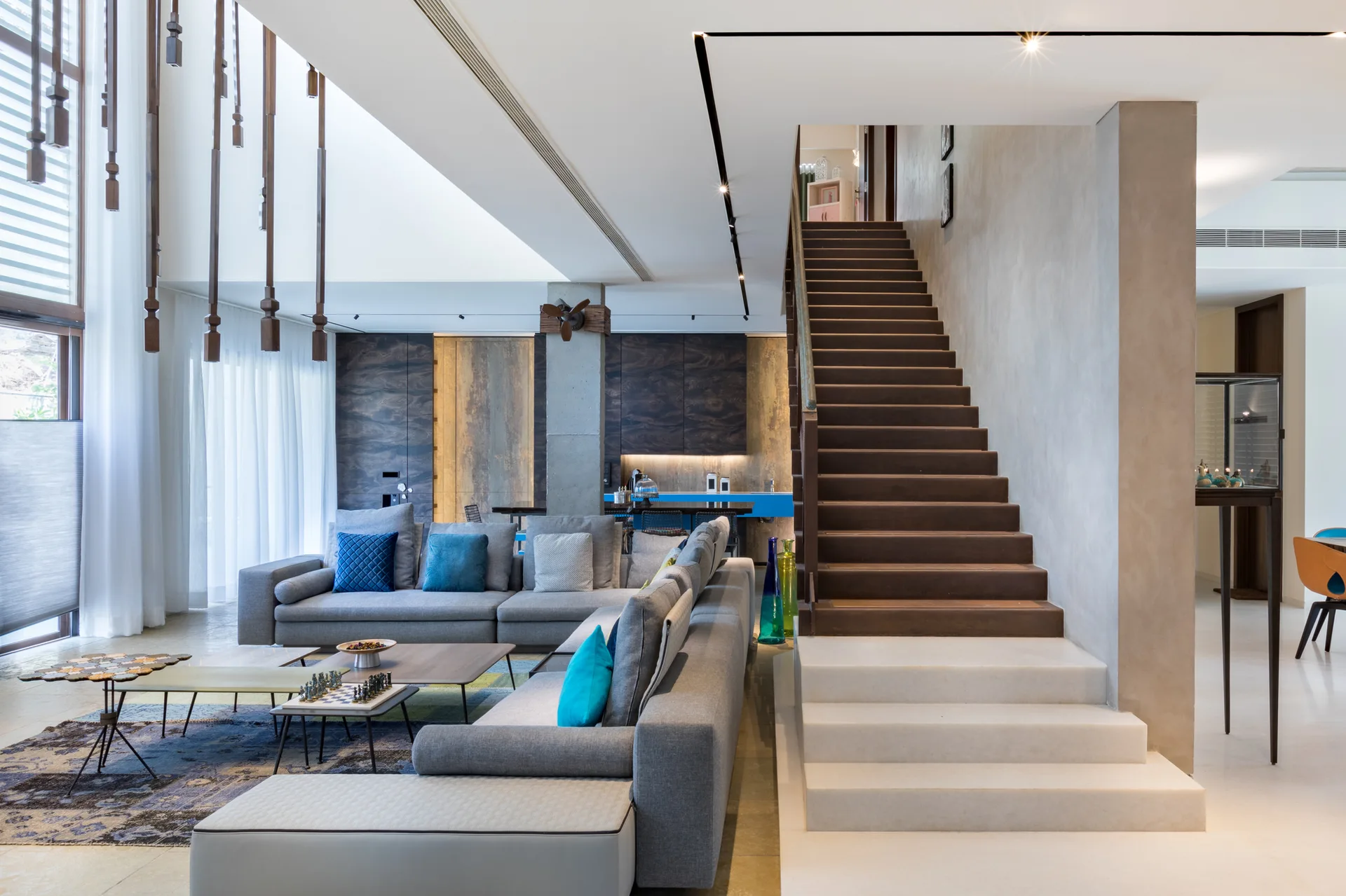A modern open-concept living room features a grey sectional sofa with blue pillows, a wood-and-stone staircase, and textured concrete walls.