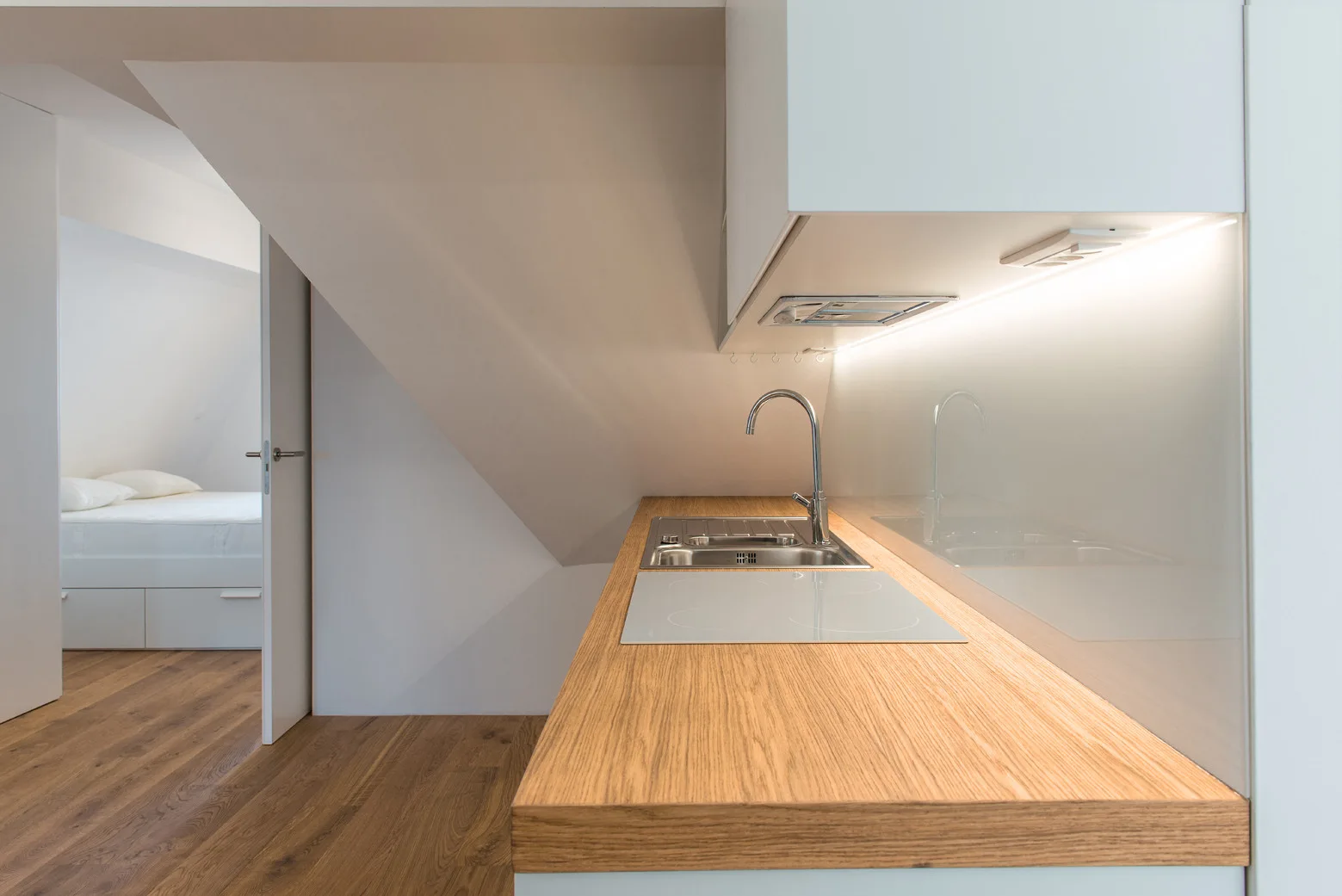 A minimalist attic kitchen features a wood-toned countertop with an inset sink beneath a sloped ceiling, looking toward an adjacent white bedroom.