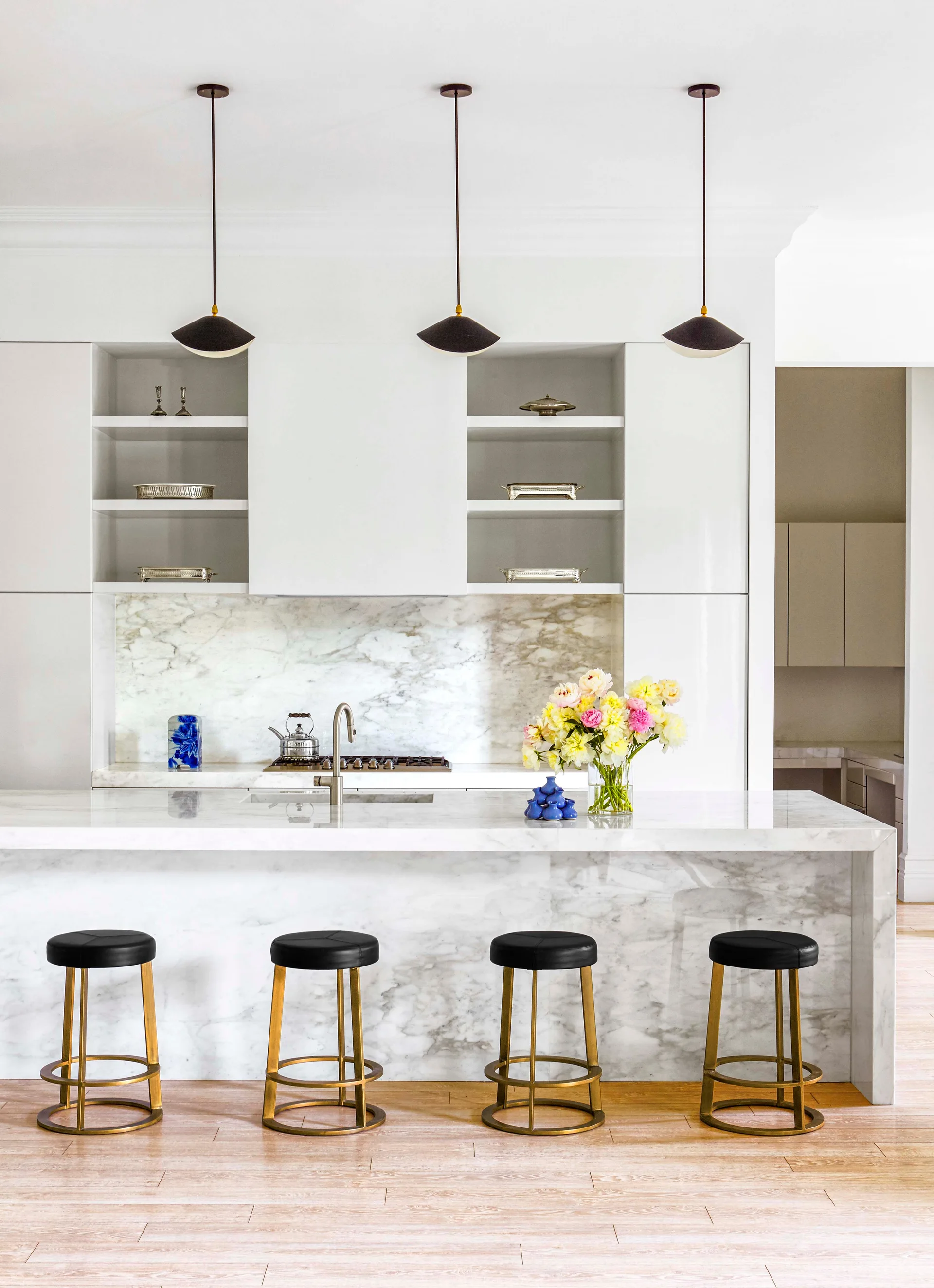 A modern kitchen featuring a white marble waterfall island with four black and gold barstools beneath three dark pendant lights.