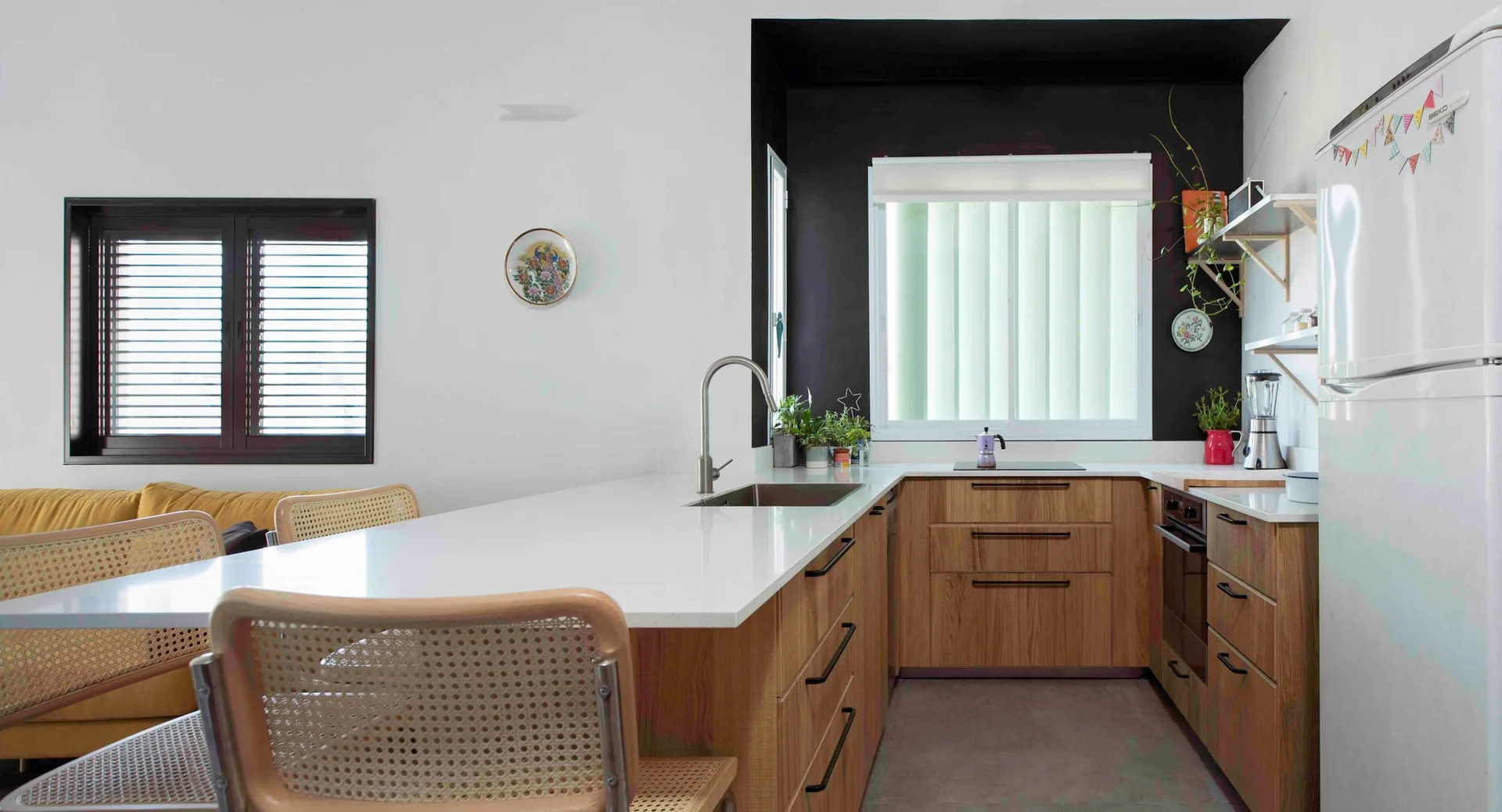 A modern kitchen with wood cabinets, white countertops, a black-painted window alcove, and light wood cane bar stools.