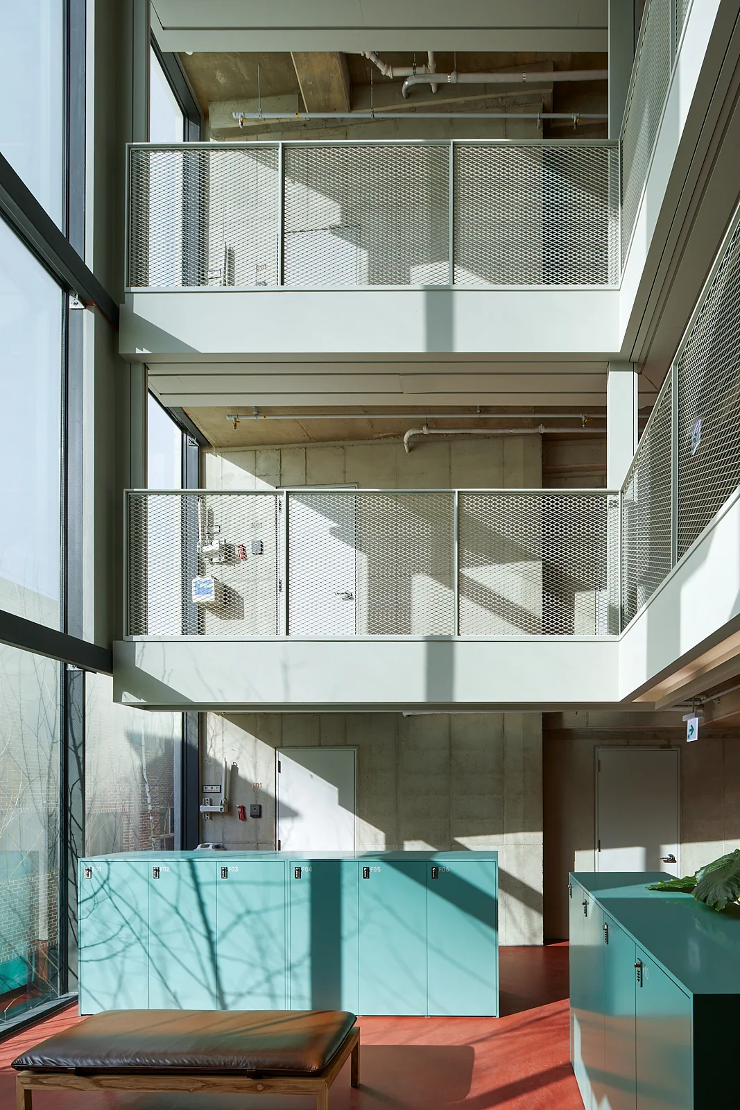 A multi-level industrial interior featuring exposed concrete walls, mesh mezzanine railings, teal lockers, and a leather bench on a red floor.