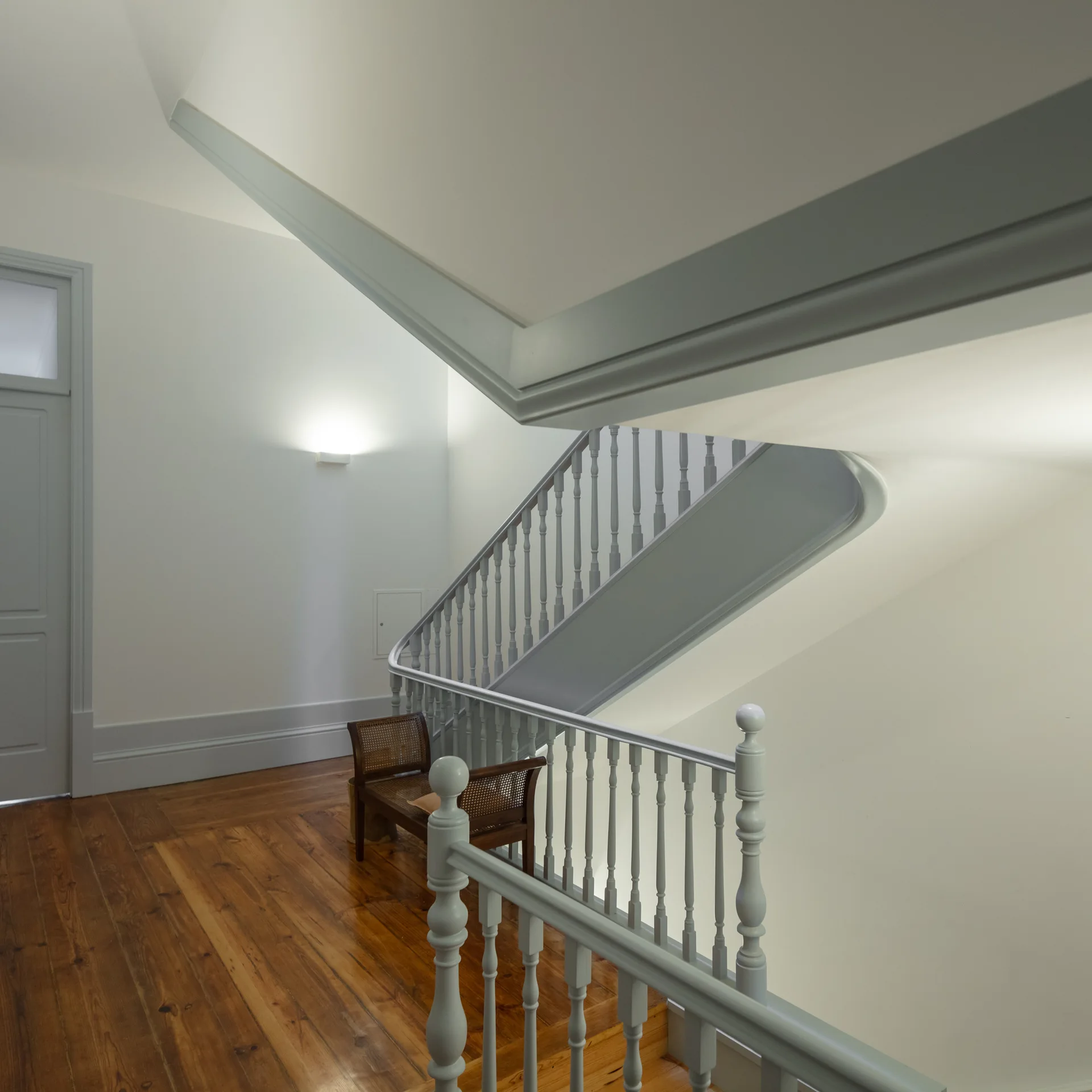 A winding white staircase features a pale grey wooden railing with vertical balusters, contrasting with warm-toned hardwood floors and a small wooden bench.