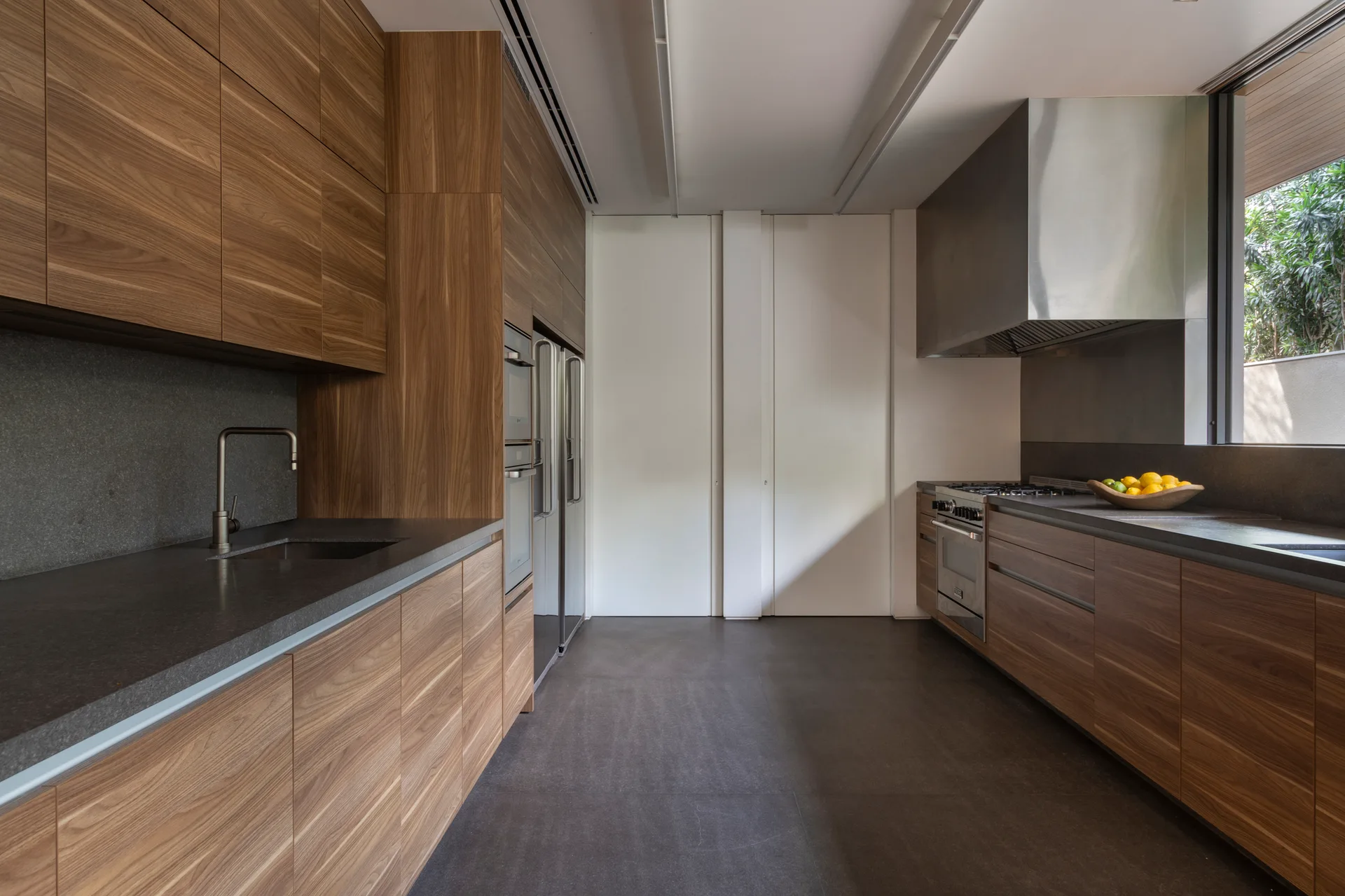A modern galley kitchen featuring flat-front wood cabinetry, dark stone countertops, a large stainless steel range hood, and matte grey floor tiles.