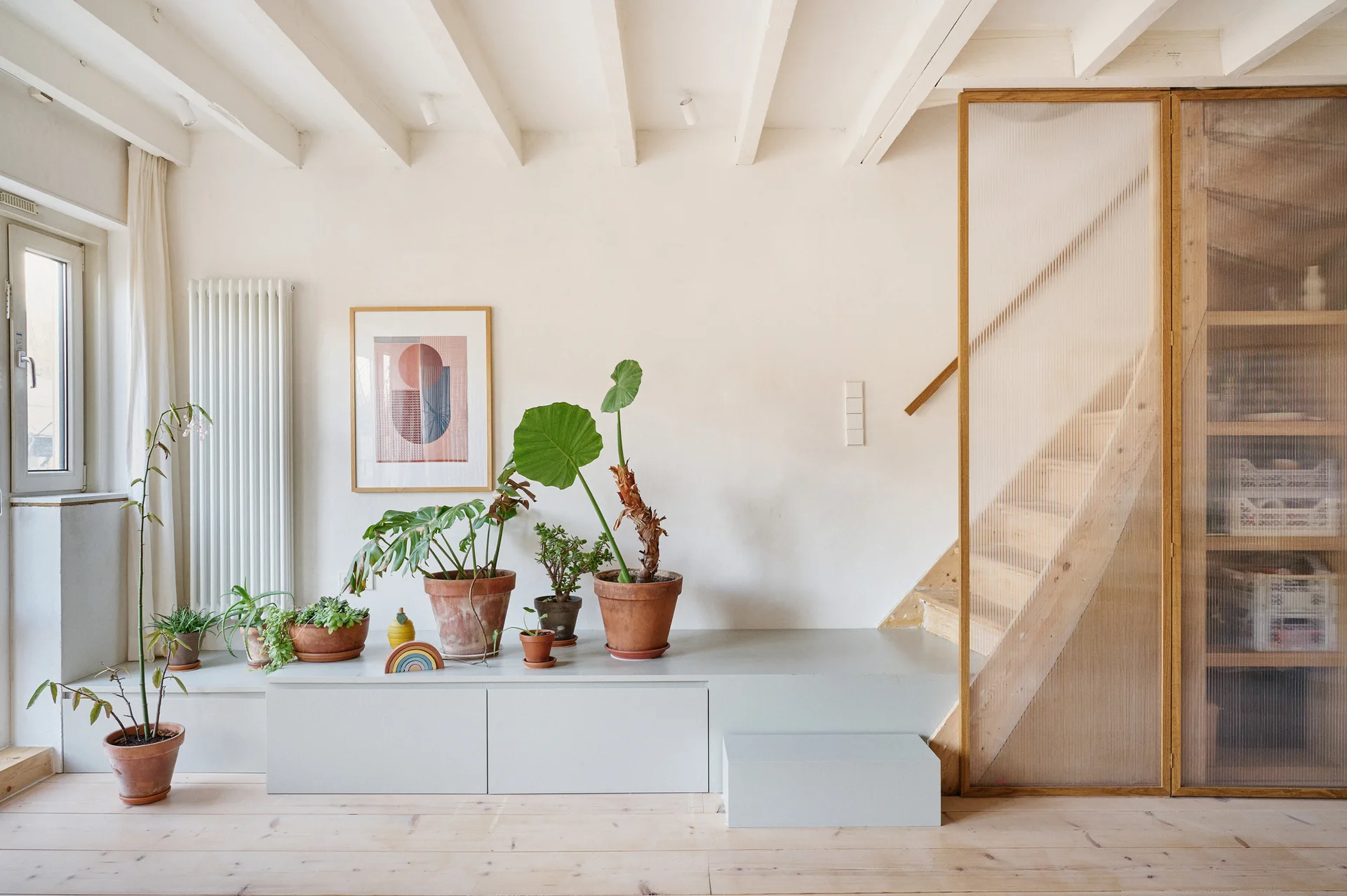 A light-filled interior shows a wooden staircase behind a fluted glass screen next to a low grey platform holding terracotta potted plants.