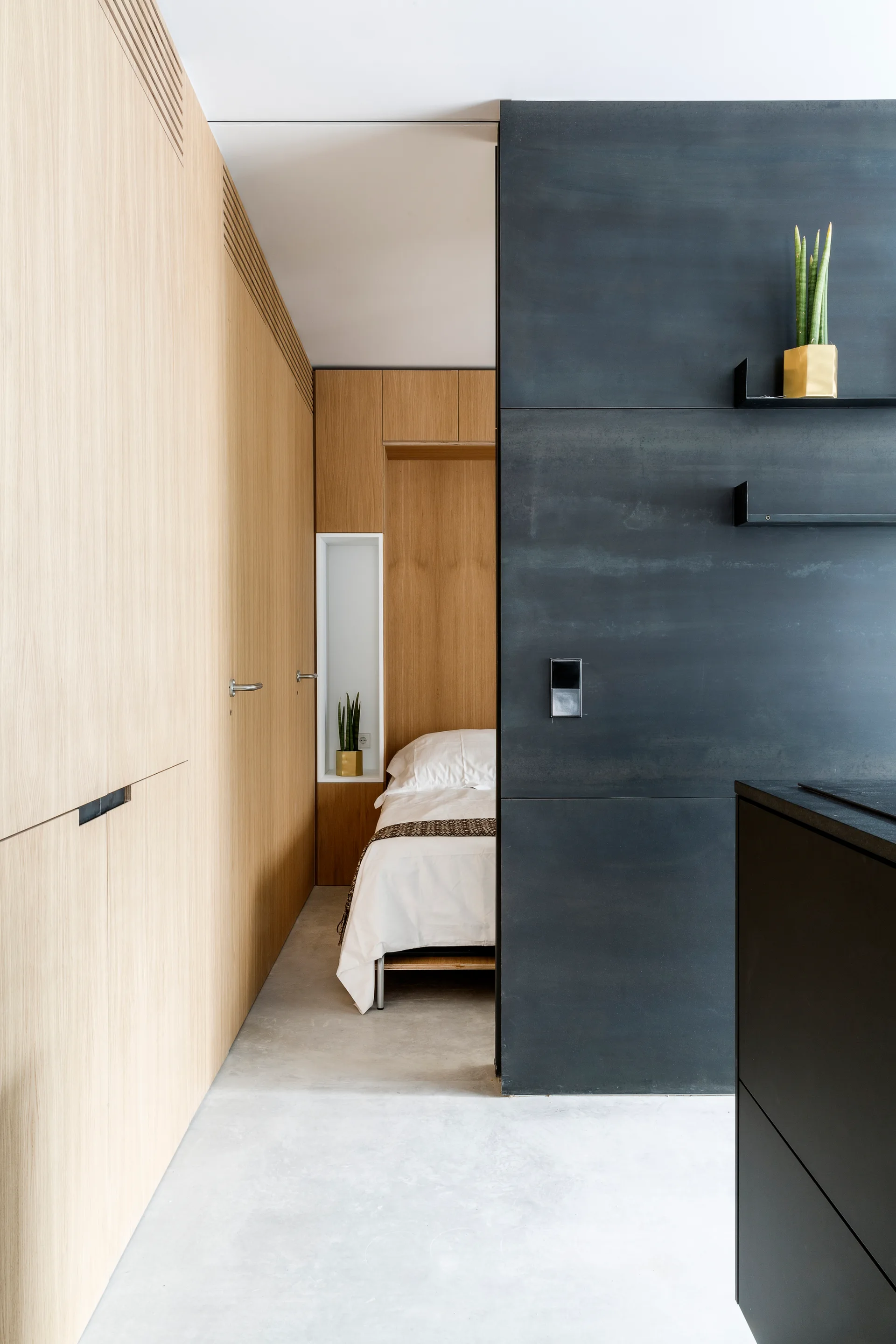 A modern bedroom view features a light wood-paneled storage wall leading to a recessed bed alcove next to a matte black accent wall.