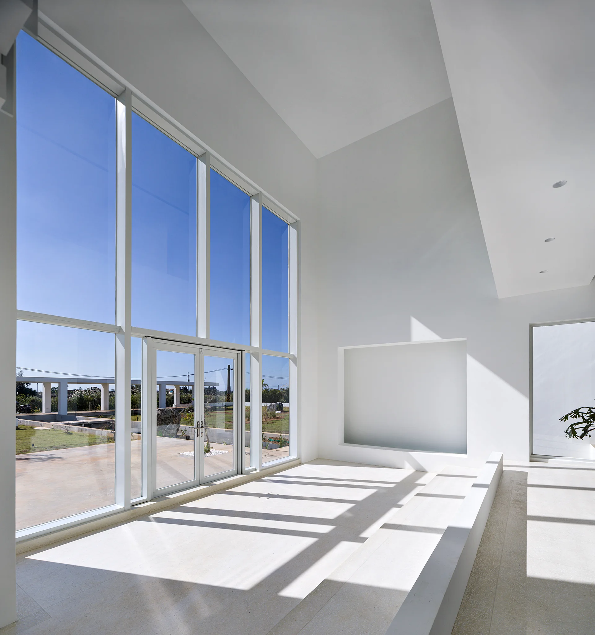 A bright minimalist white room features a massive floor-to-ceiling glass window wall, a stepped stone floor, and a recessed wall niche.