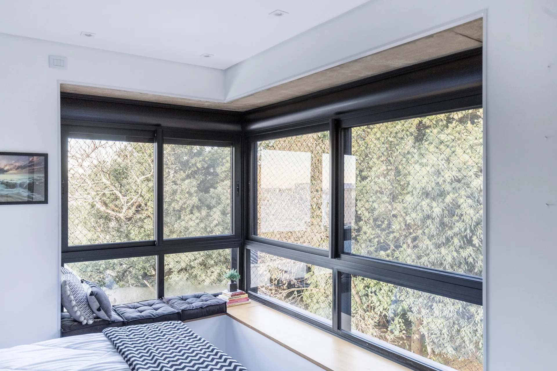 A minimalist bedroom corner featuring a built-in light wood window seat with dark cushions beneath large black-framed corner windows.
