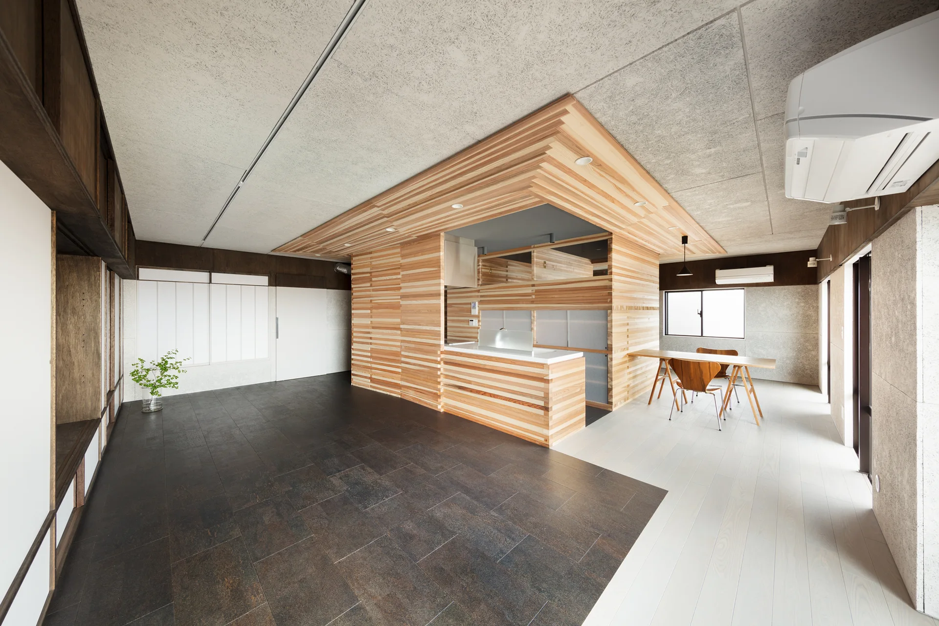 A minimalist open-plan kitchen and dining area featuring a horizontal slatted wood central structure, dark tile flooring, and a wooden dining table.
