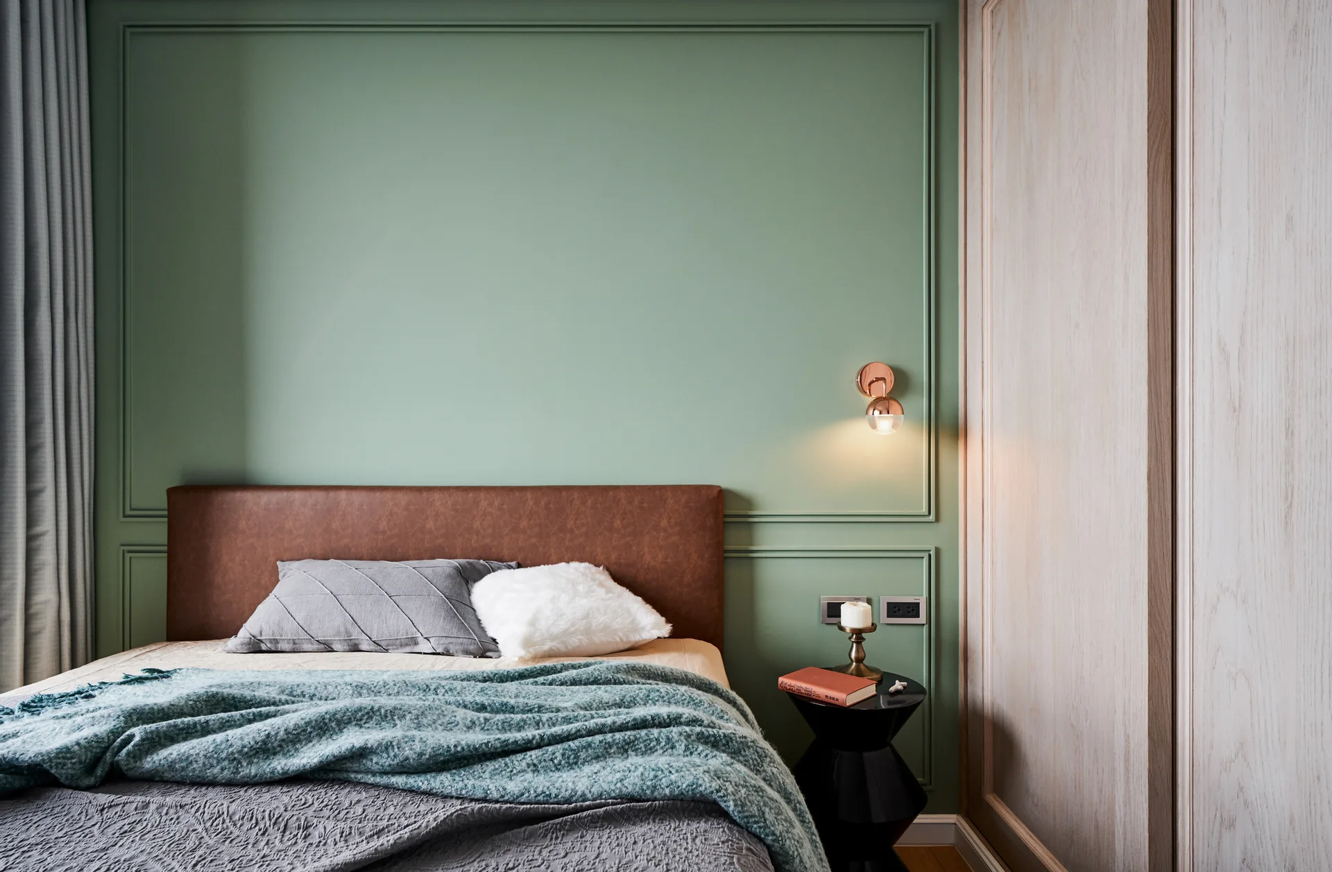 A bedroom featuring a brown leather headboard against a sage green paneled wall, paired with a black pedestal side table and a glowing copper wall sconce.