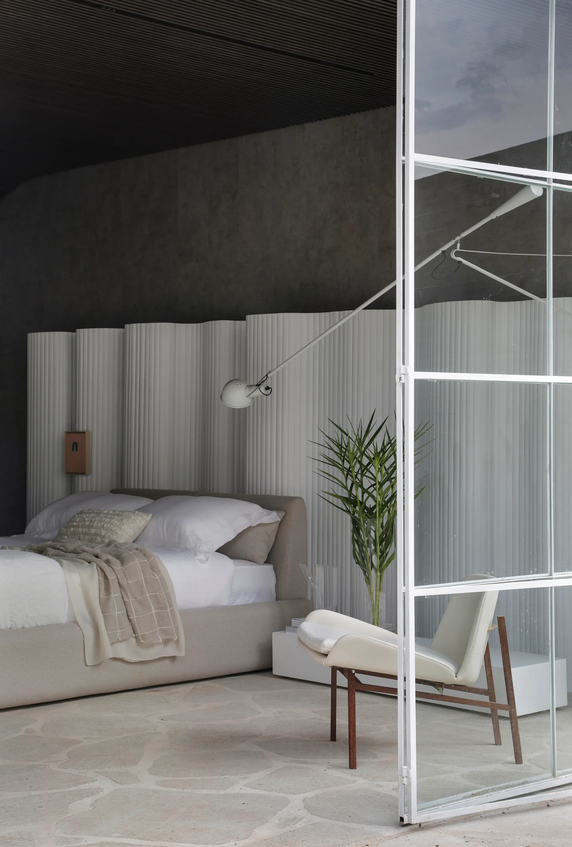 A modern bedroom features a low upholstered bed against a white curved fluted partition, next to a light-colored armchair and a large grid window.