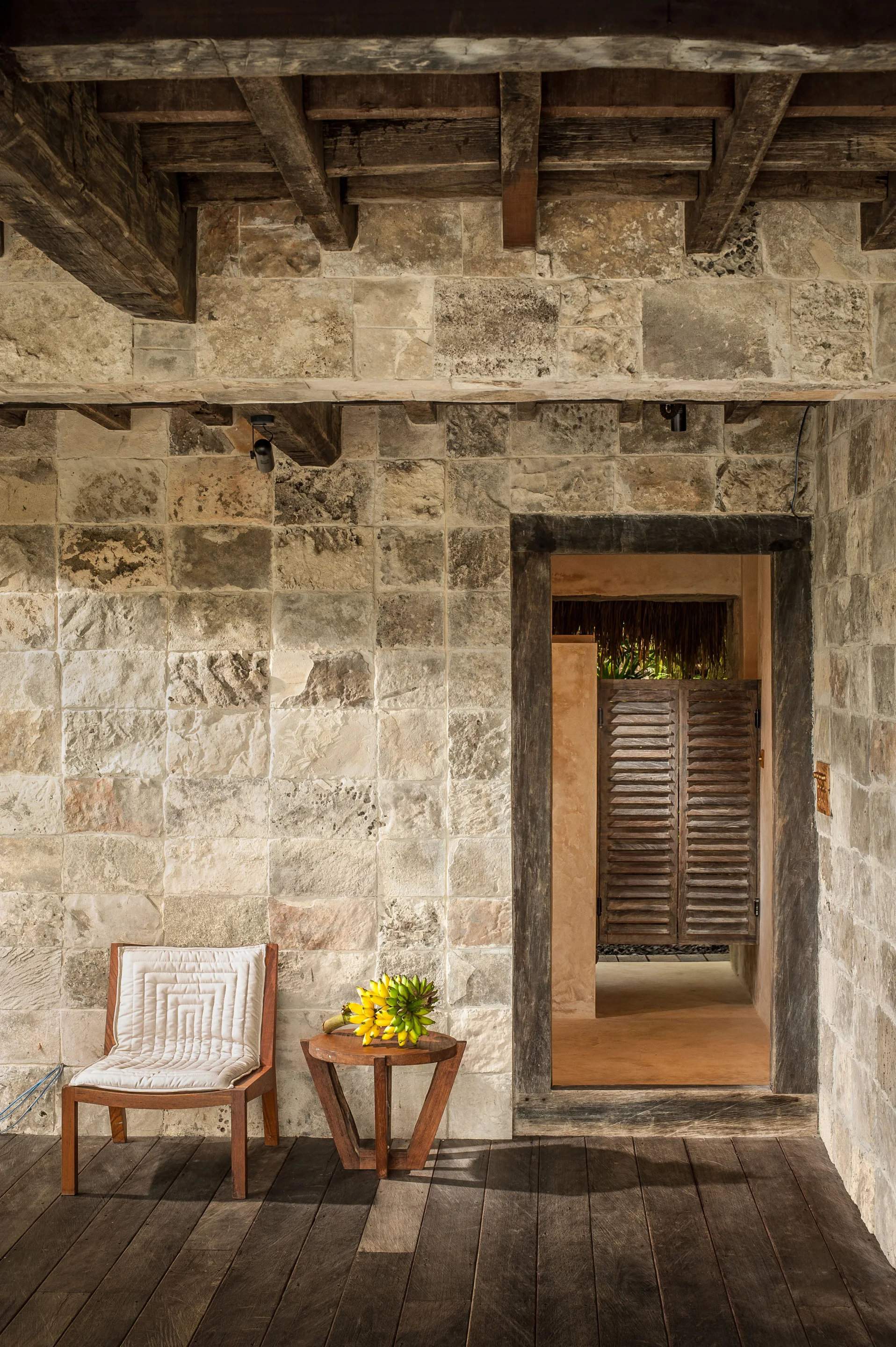 A rustic patio features a textured stone block wall, exposed dark wood ceiling beams, a wood-framed chair with a white cushion, and a side table.
