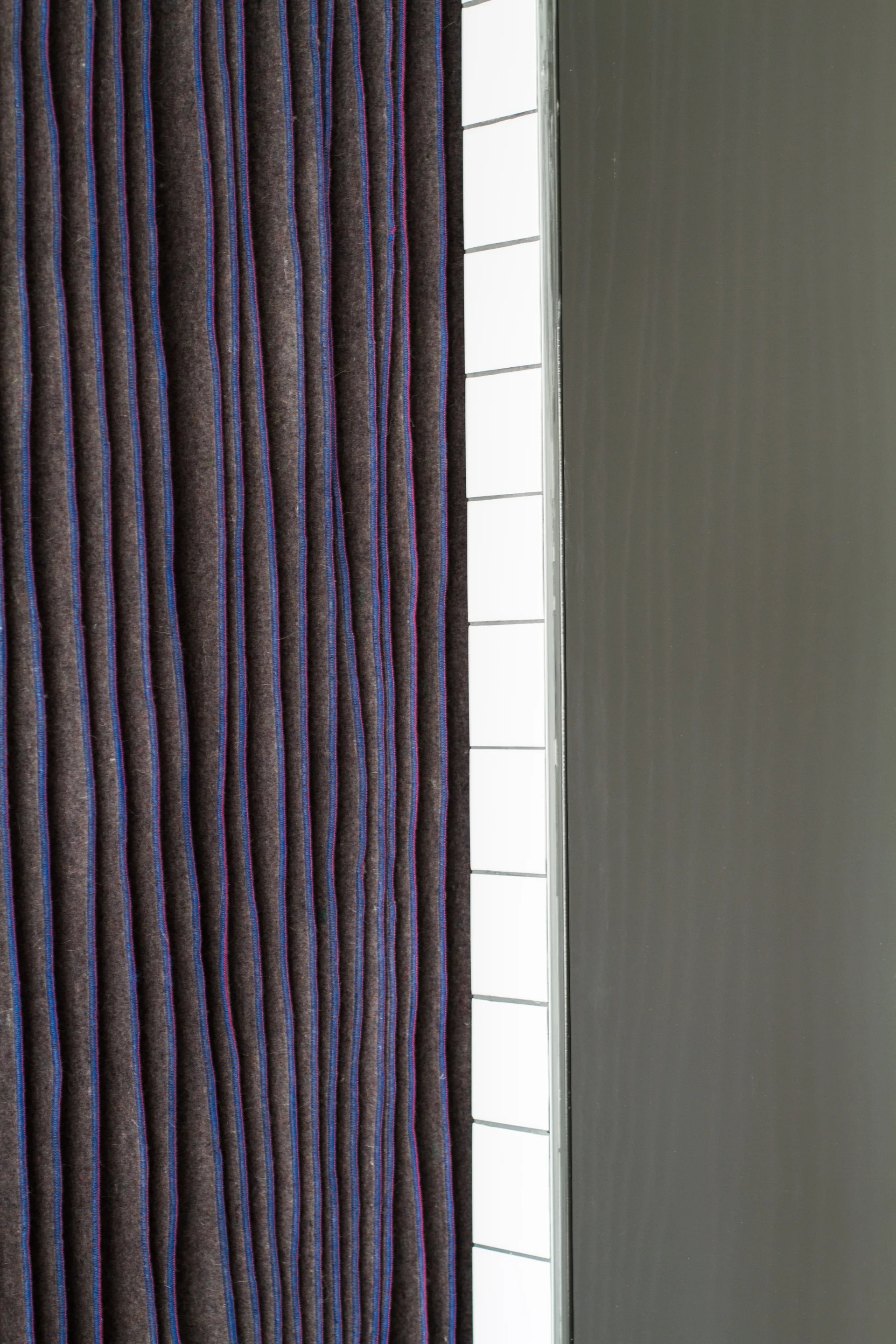 A close-up view of layered dark grey felt panels with blue edges positioned next to a vertical strip of white square tiles and a solid grey surface.