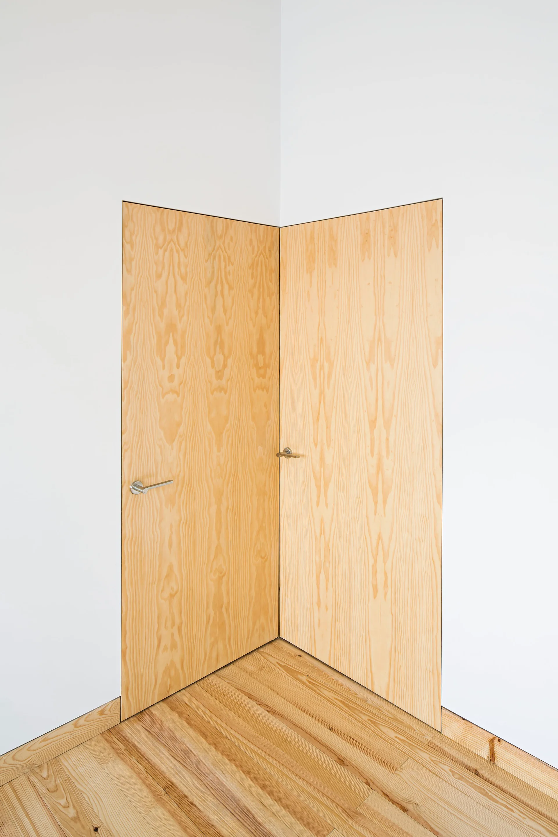 Two frameless wood veneer doors are installed flush against a white corner wall above light wood flooring.
