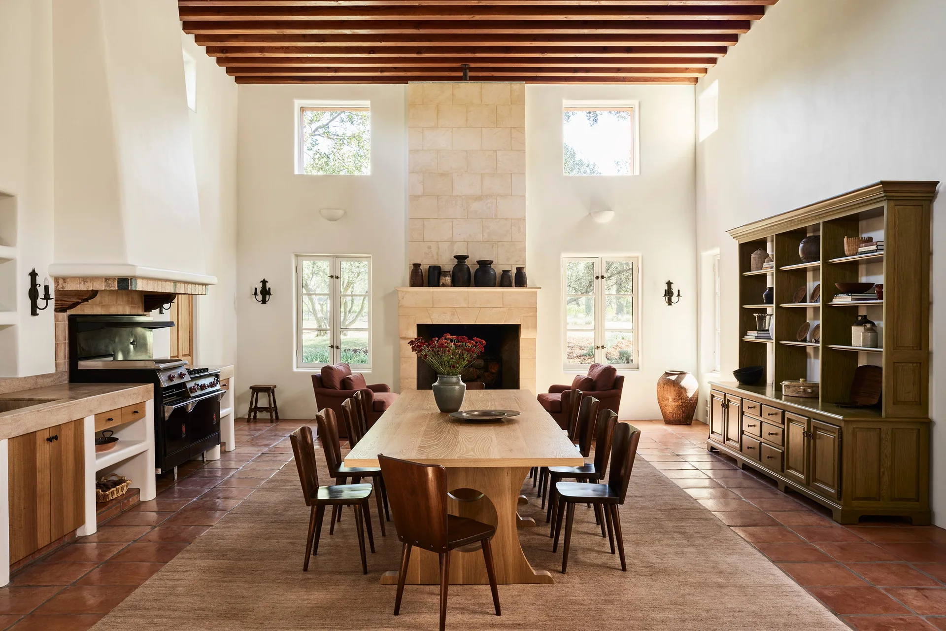 A rustic dining room features a long wooden table, terracotta tile flooring, exposed ceiling beams, and a monumental stone fireplace flanked by windows.