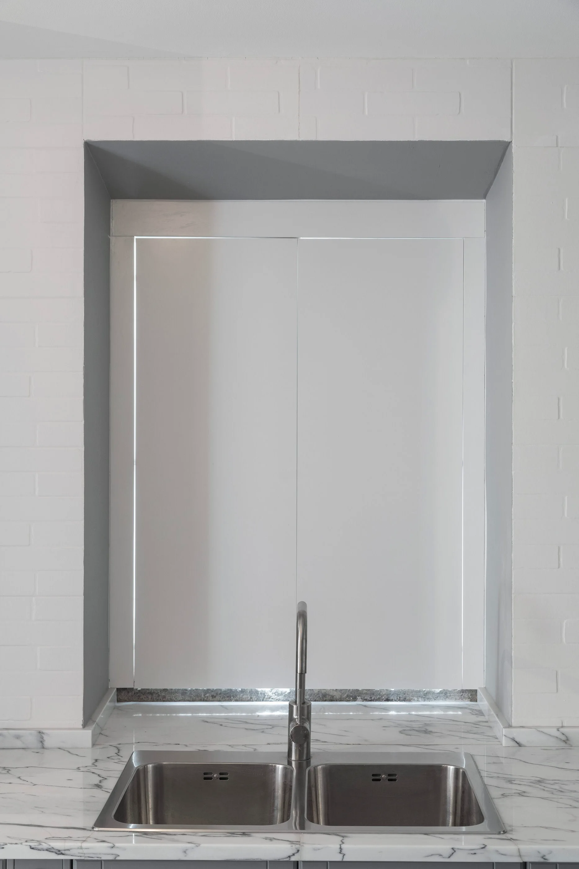 A stainless steel double kitchen sink is set in a white veined marble countertop below a deep recessed window niche with white panels.