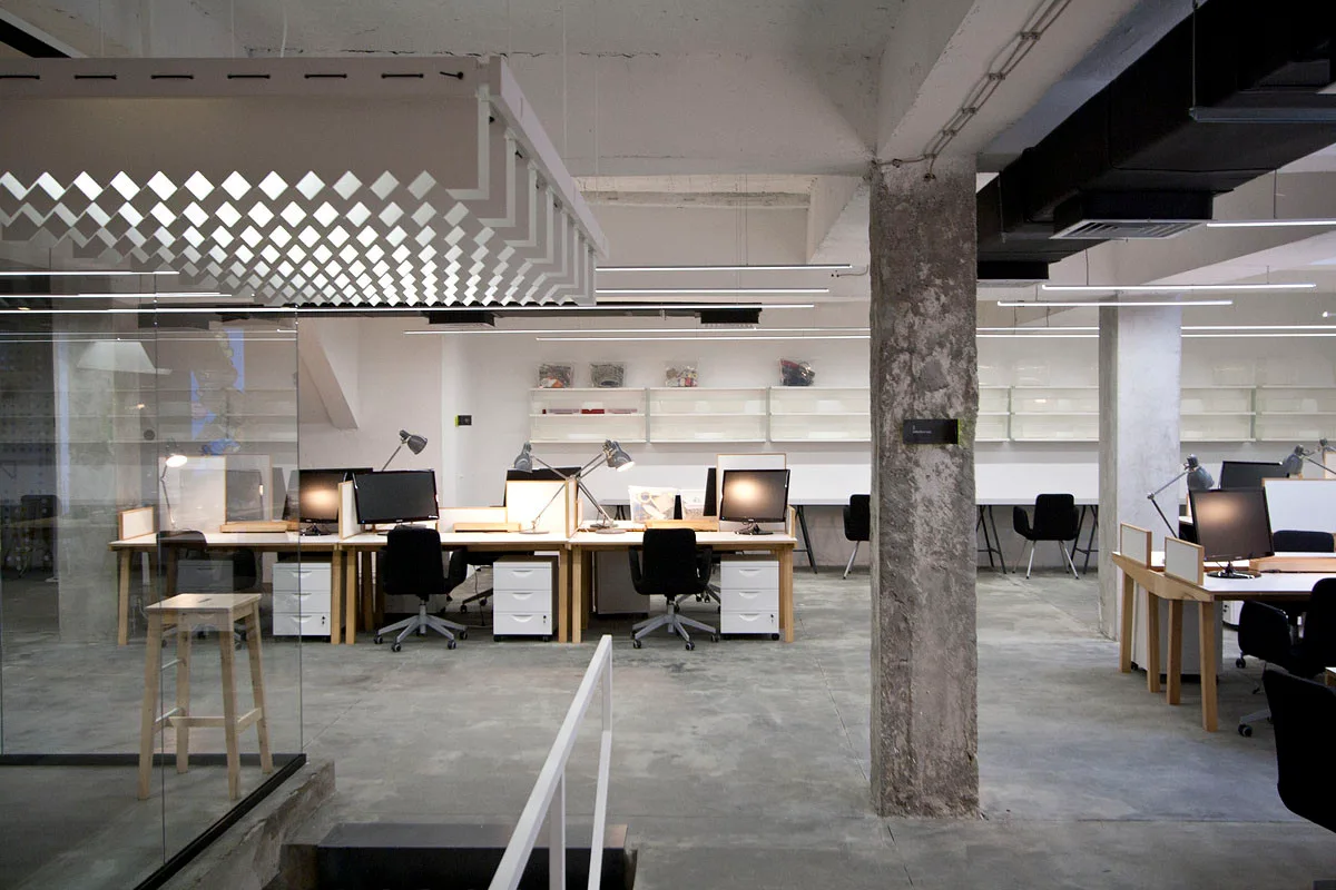 An open-plan industrial office features long wood desks, black rolling chairs, a raw concrete pillar, and suspended linear light fixtures.
