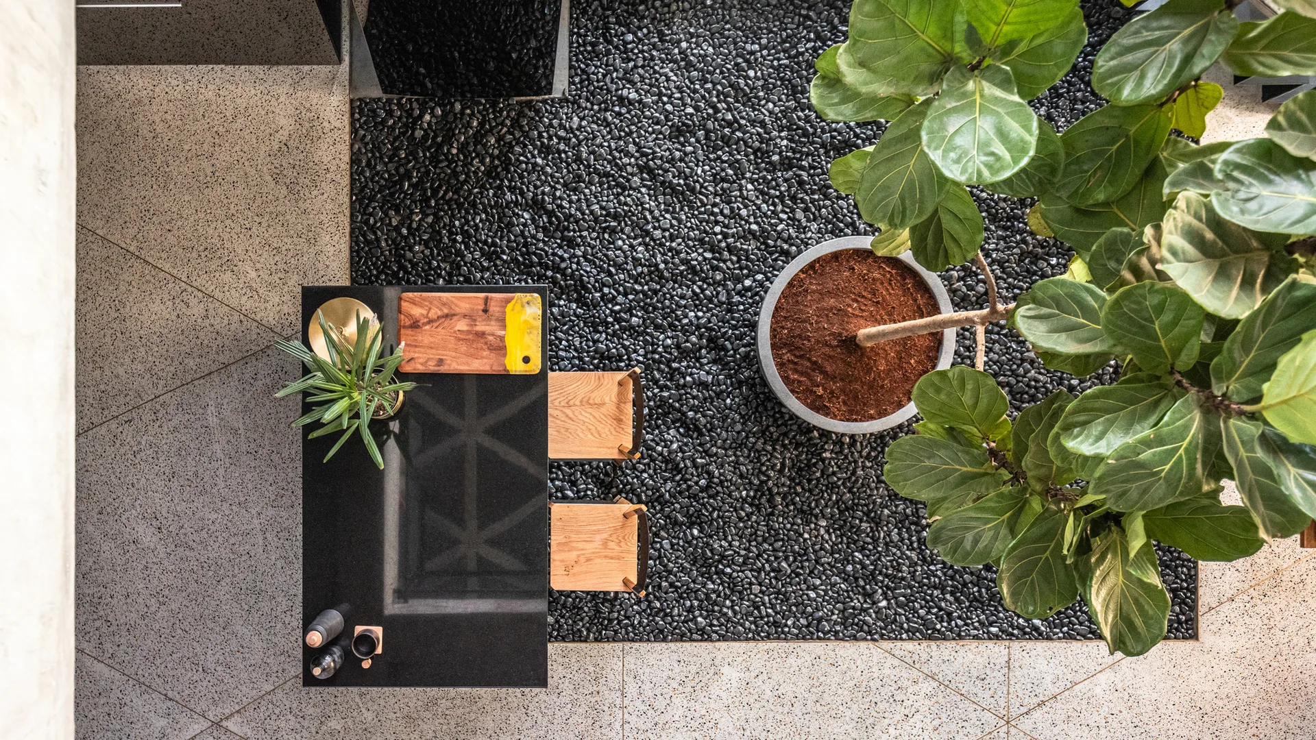 An overhead view of a black dining table and wooden chairs set upon a dark pebble floor insert surrounded by speckled terrazzo tile.