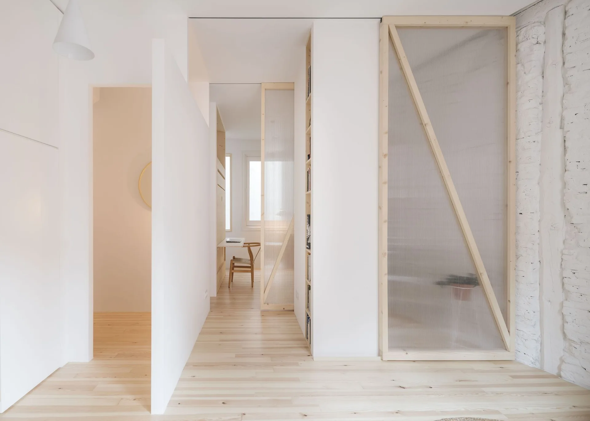 A minimalist white hallway featuring light wood flooring, built-in vertical shelving, and wooden-framed translucent fluted partition walls.