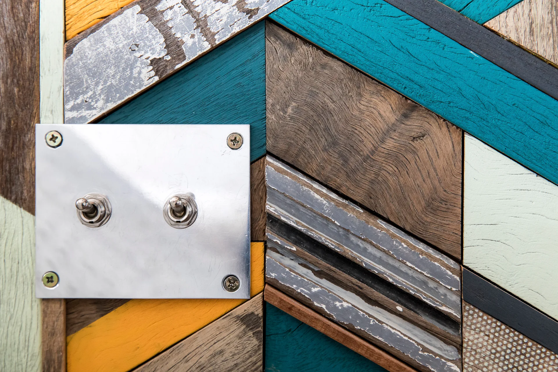 A brushed metal dual toggle light switch plate is mounted on a multicolored geometric wood plank accent wall.