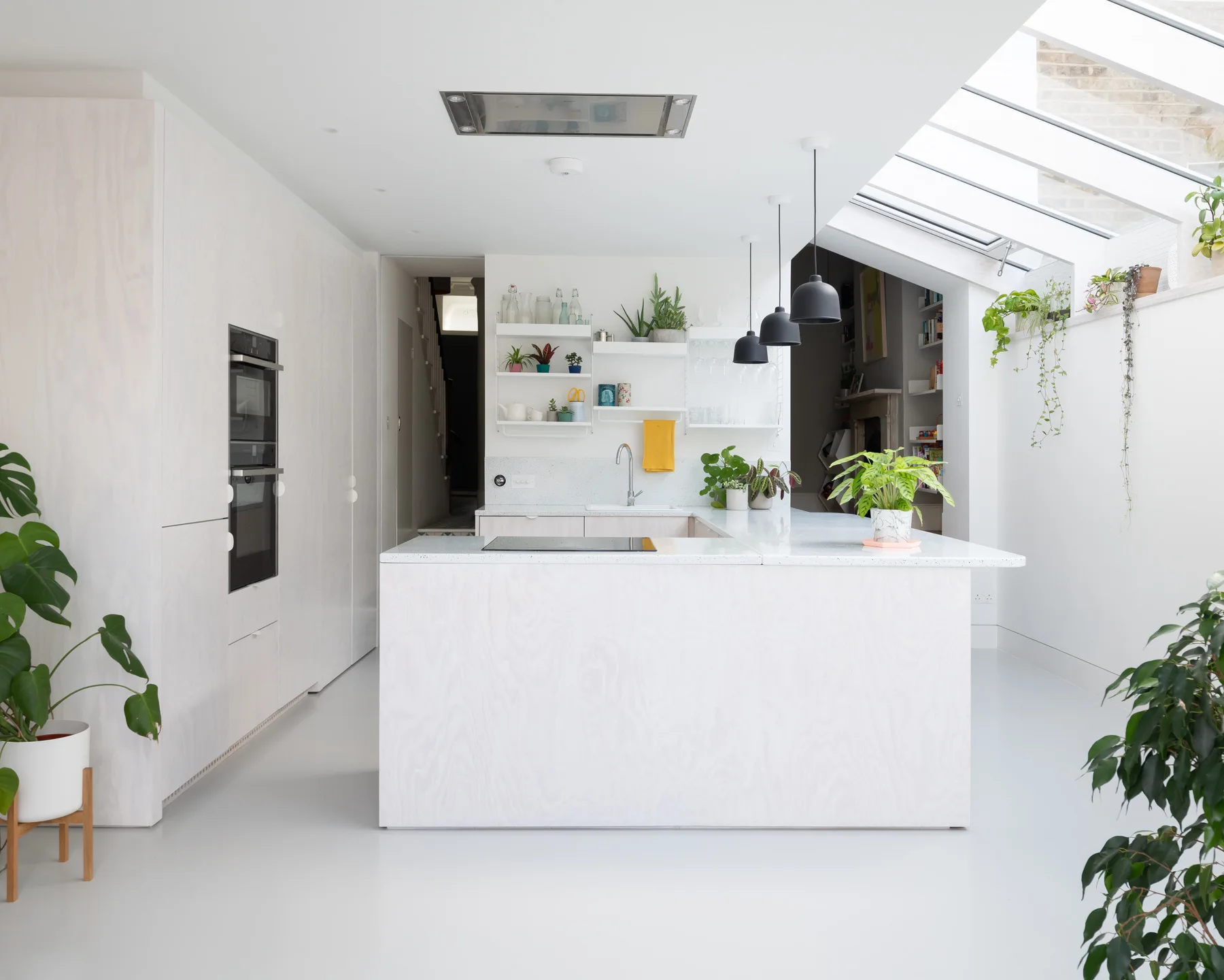 A minimalist white kitchen features a large central island, light wood-grain cabinets, black pendant lights, and a sloped glass skylight ceiling.