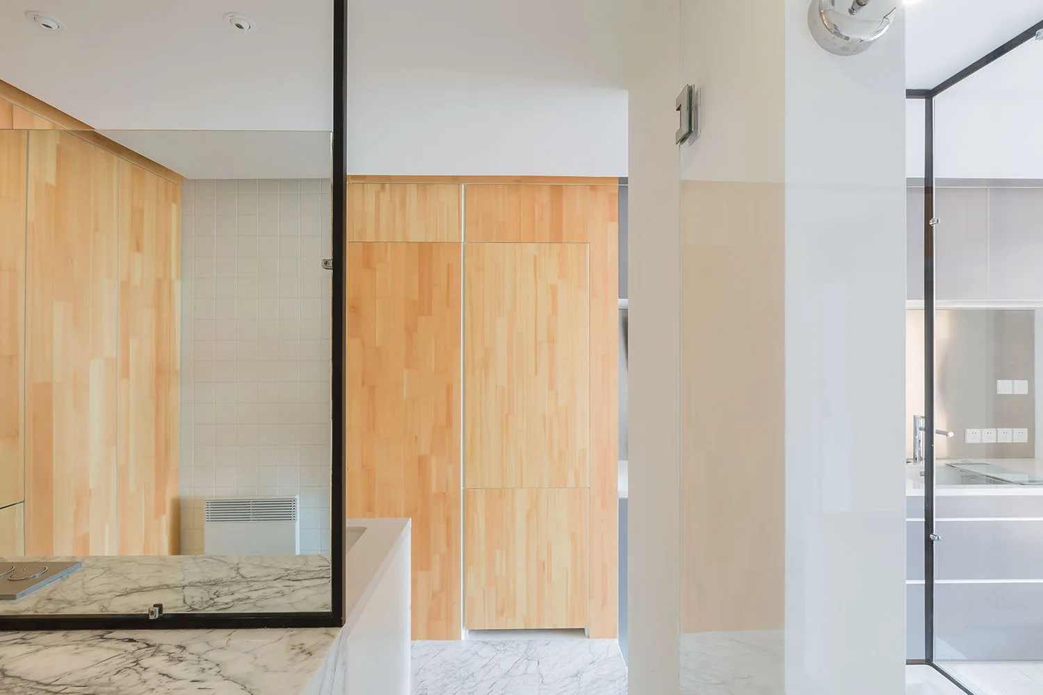 A minimalist bathroom features a veined white marble vanity, a large mirror, seamless light wood wall paneling, and a glass-enclosed shower.