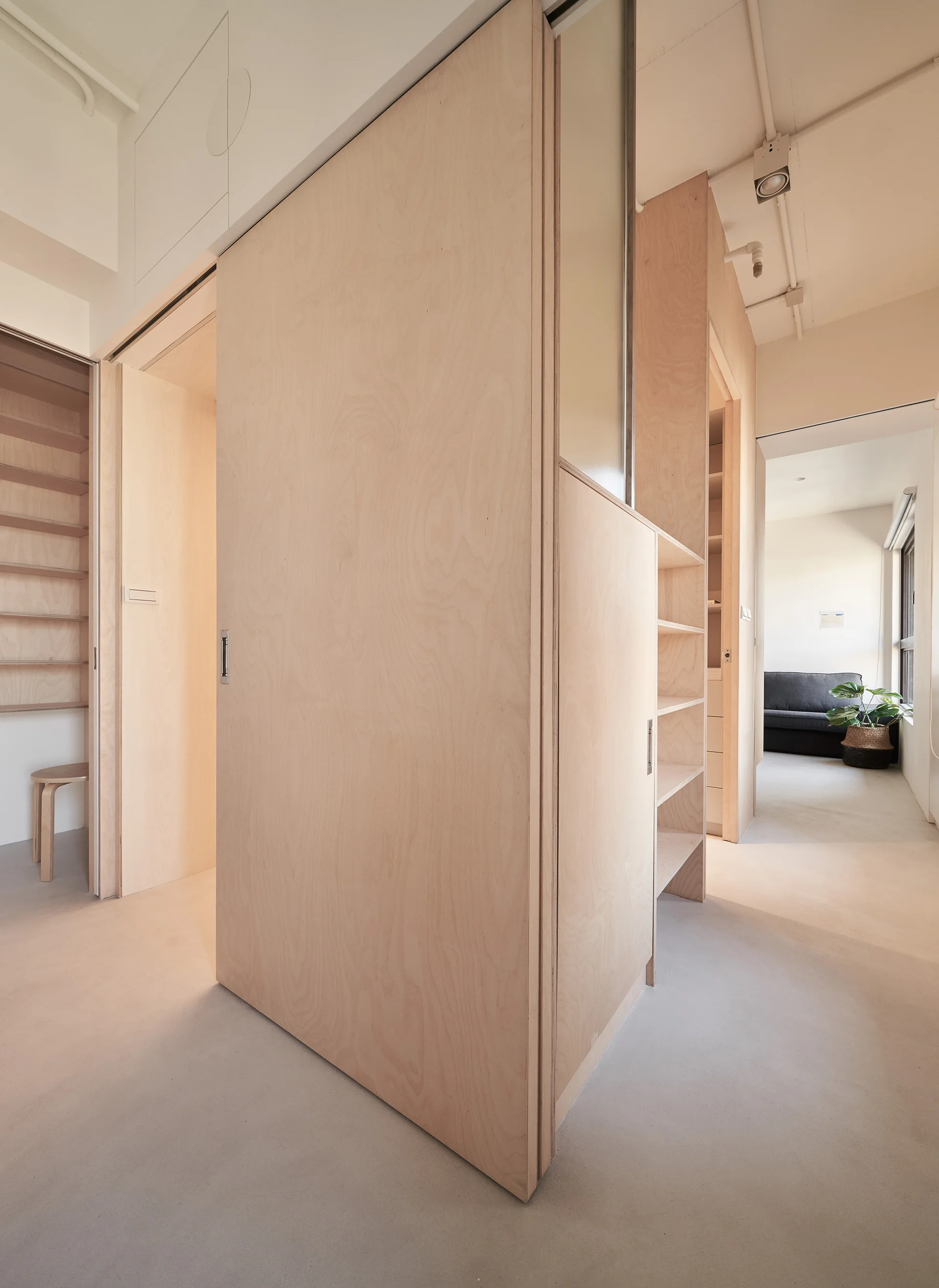 Custom light-colored plywood partition walls and built-in shelving units frame a hallway with smooth grey floors leading to a bright living area.