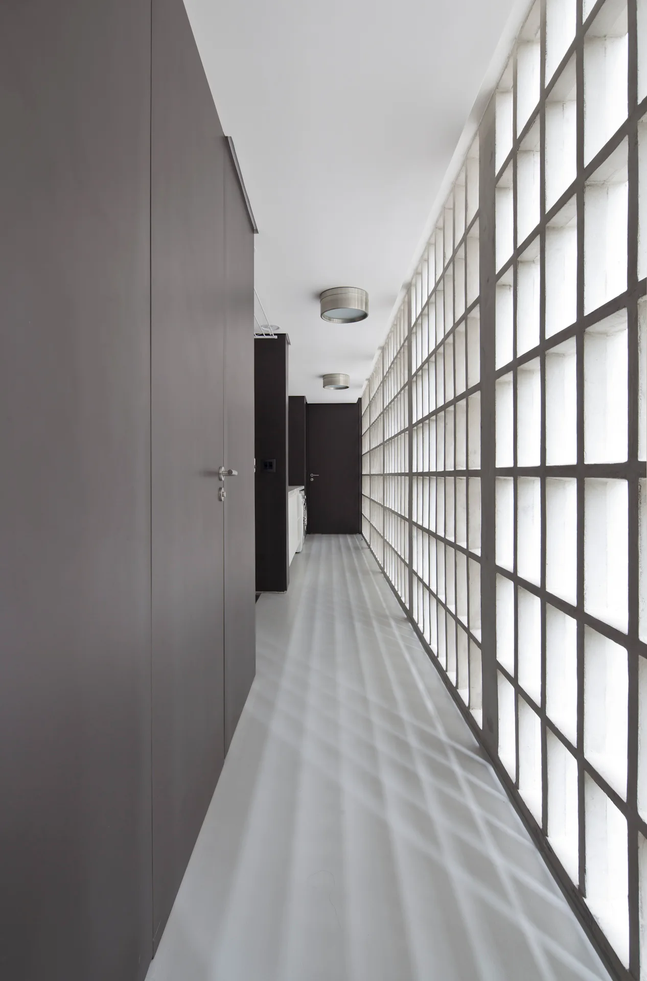 A long minimalist hallway with a floor-to-ceiling glass block wall casting grid shadows across a light grey floor opposite dark flat cabinet doors.