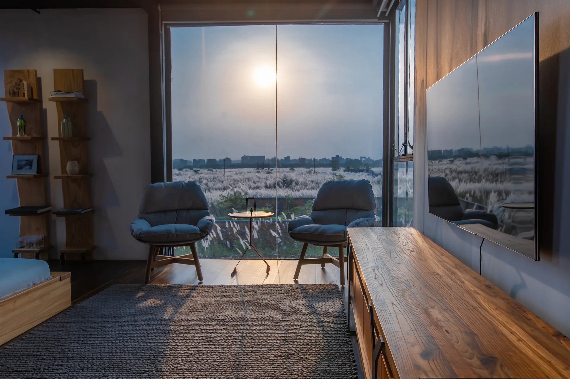 Two gray upholstered lounge chairs face a large glass window overlooking a grassy field at sunset, next to a long wooden media console.