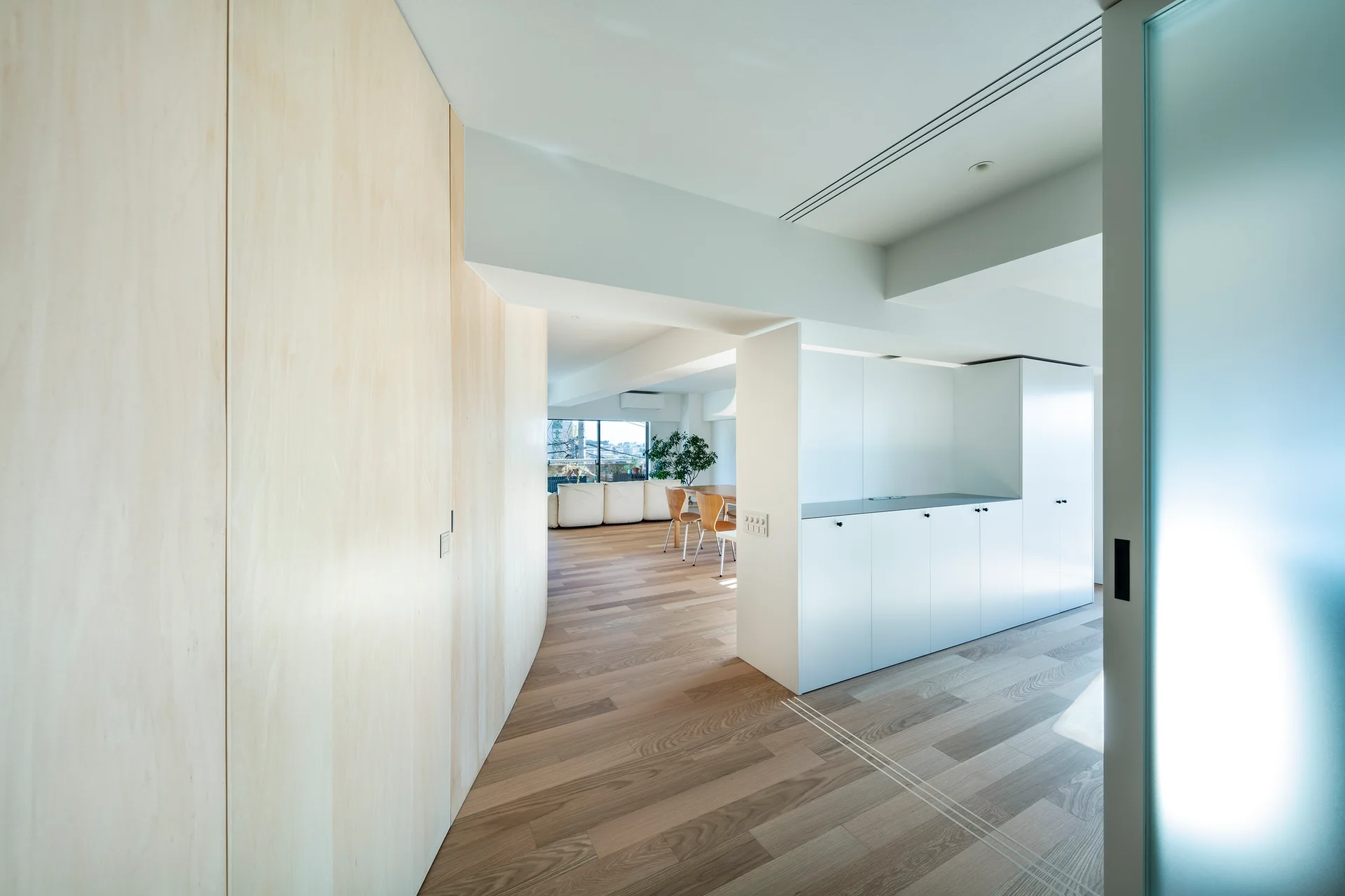 A minimalist hallway features curved light wood wall paneling and matte white built-in storage cabinets leading to an open living and dining area.