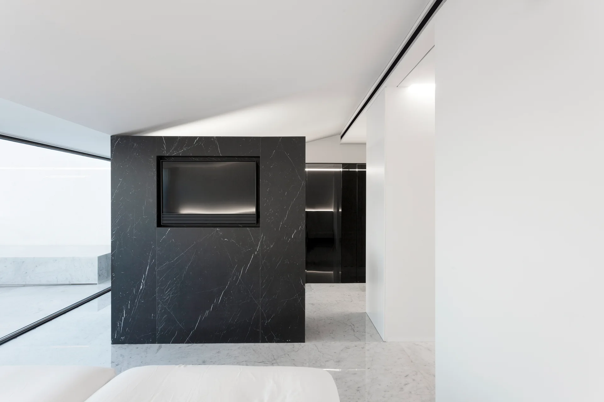 A minimalist living room displays a black marble partition wall with a recessed screen, set on veined white marble flooring under a white ceiling.