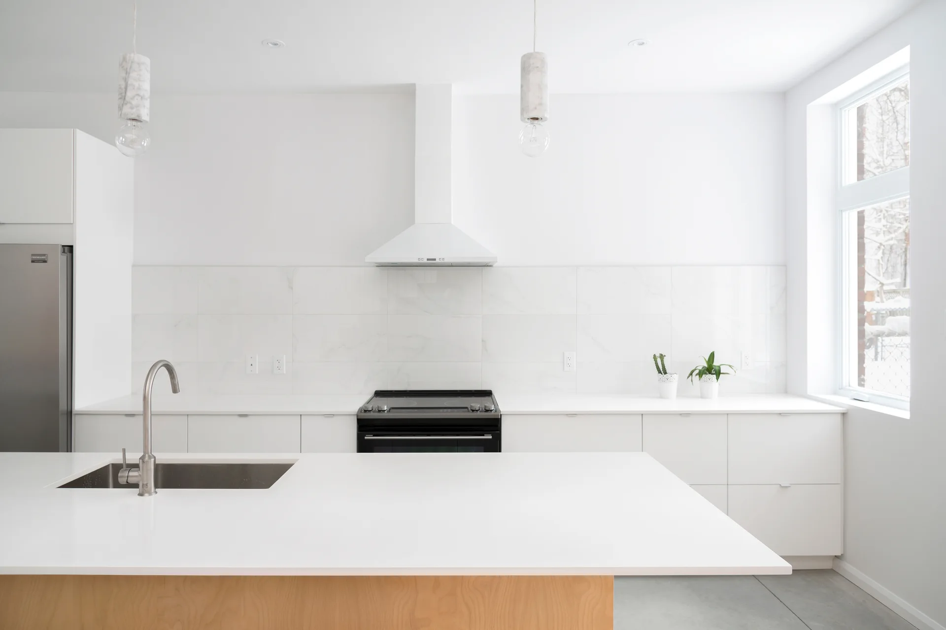 A minimalist white kitchen featuring a large white island with a wood base, a tiled backsplash, and stone cylinder pendant lights.