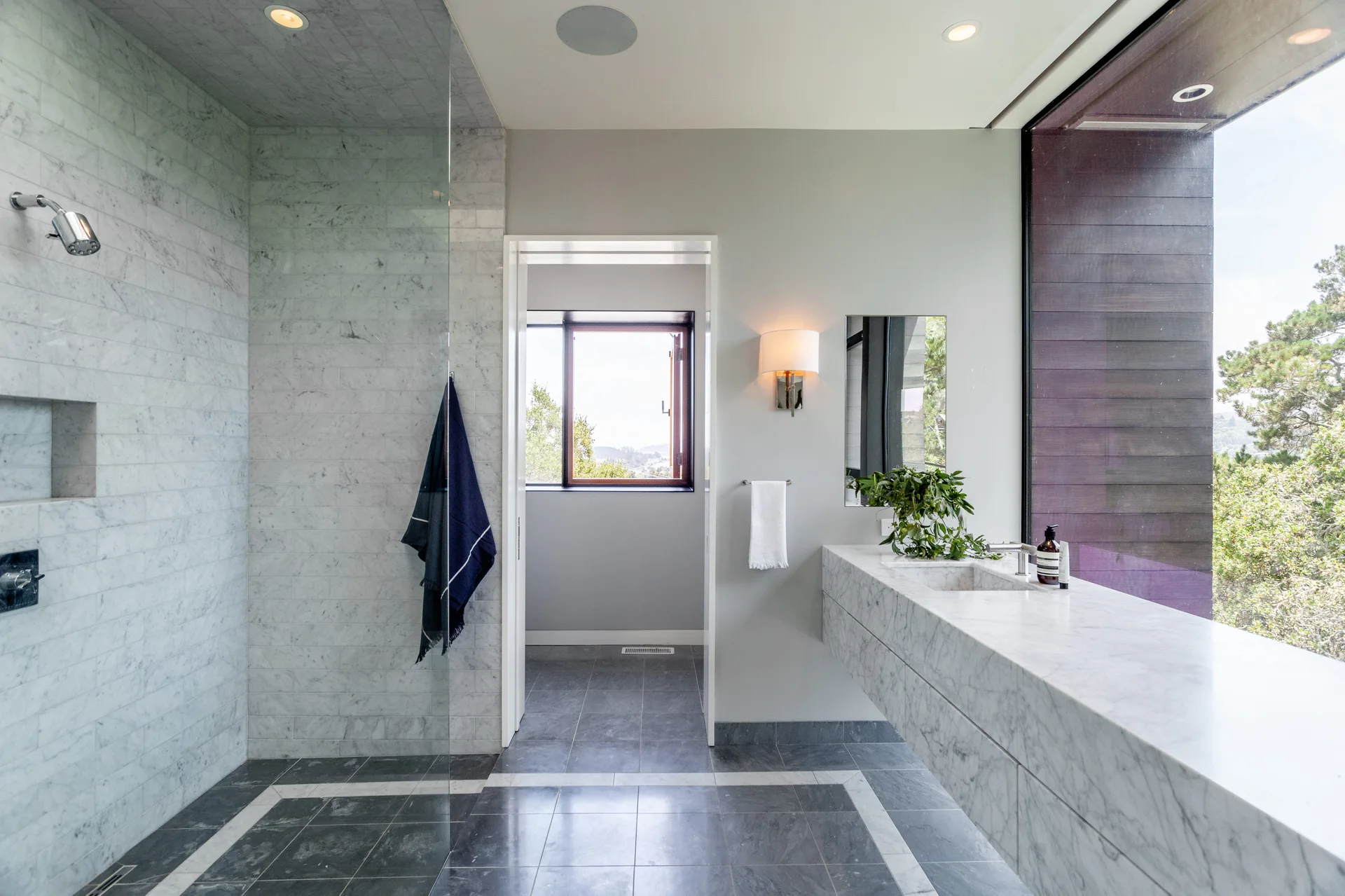 A contemporary bathroom features a long floating marble vanity with a sink, a glass-enclosed shower with marble tiles, and a large window.