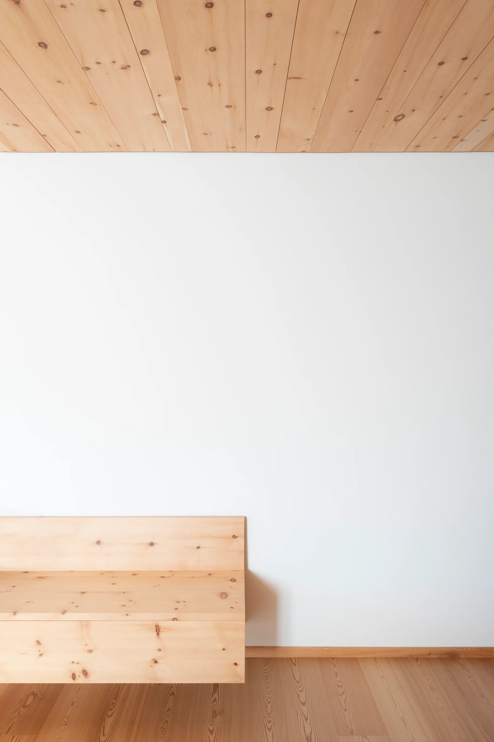 A minimalist room features a built-in light wood bench against a plain white wall with a matching wood plank ceiling and wood floor.