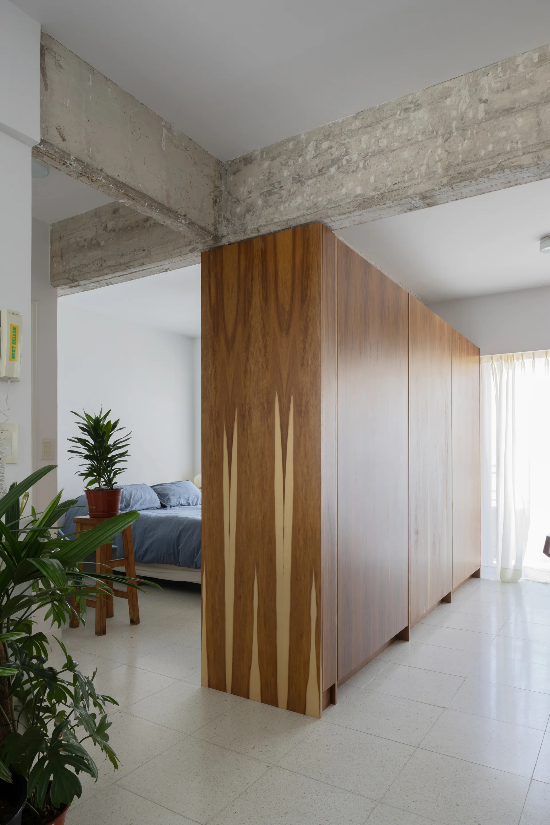 A large wood veneer partition wall divides an apartment, showing a bedroom with blue bedding next to raw concrete ceiling beams.