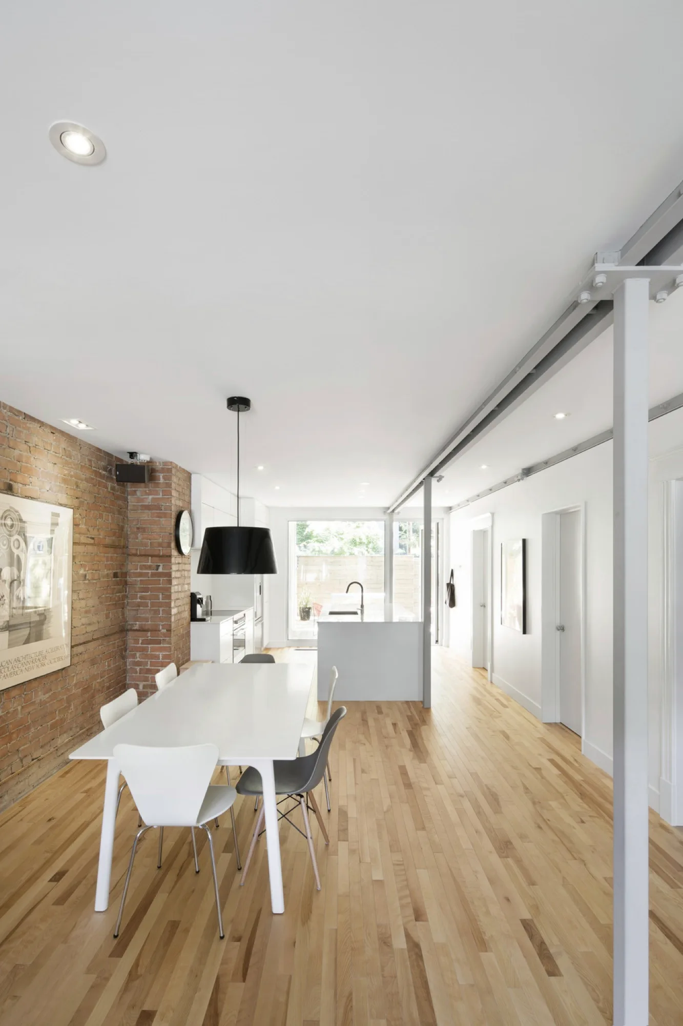 A long modern dining area features a white dining table with chairs next to an exposed brick wall, leading to a white open-plan kitchen.