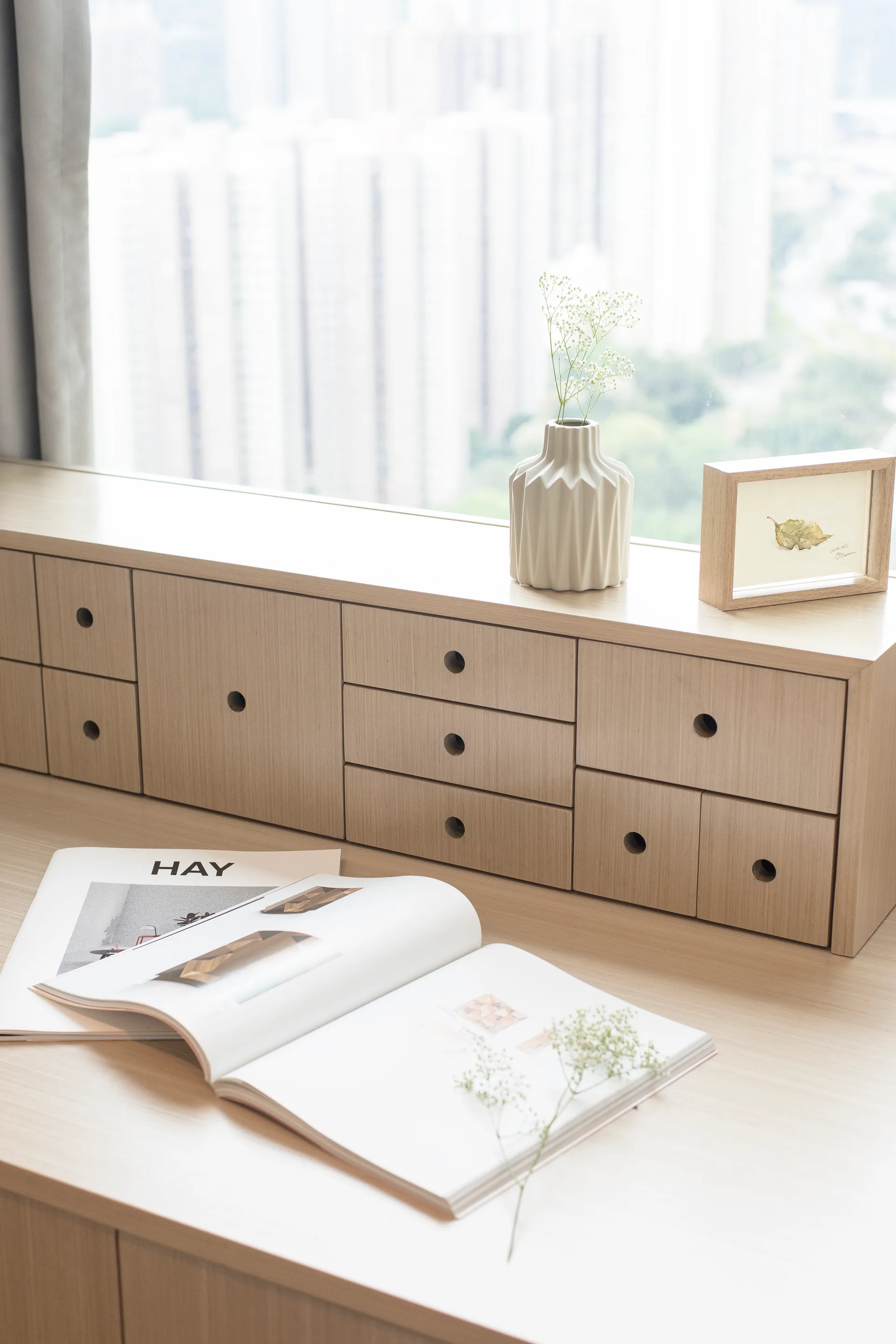 A light oak desk organizer with small drawers sits by a window next to a ceramic vase, picture frame, and open magazines.