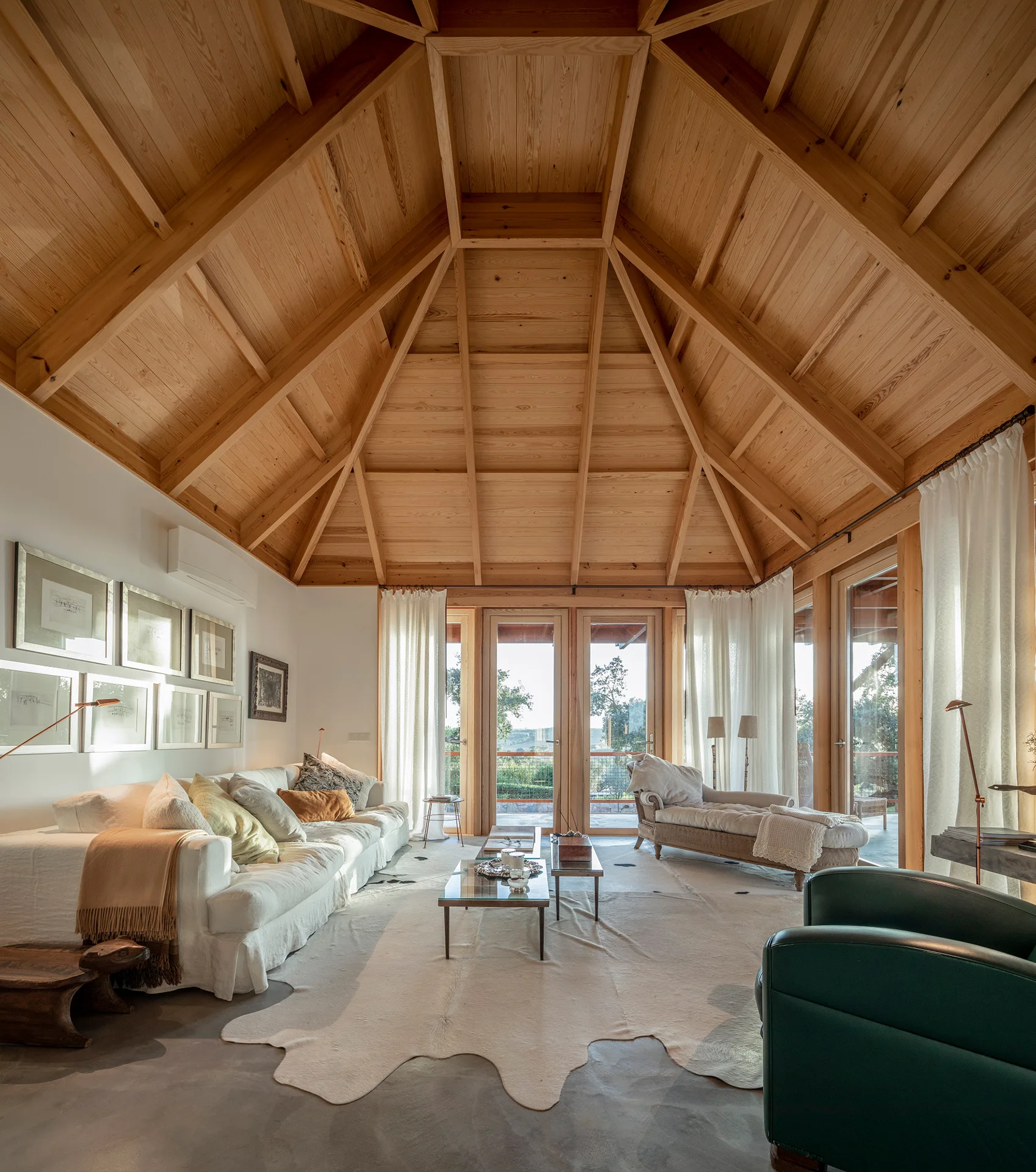 A living room features a vaulted wood ceiling, white sofa, a matching chaise lounge, sheer white curtains, and a cowhide rug on concrete flooring.