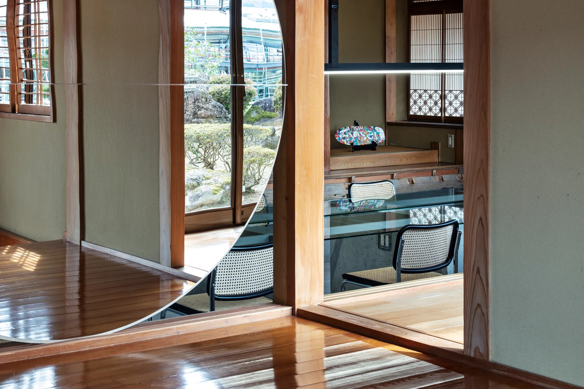 A Japanese style room featuring polished wood floors, a glass top table with cane chairs, and a large circular mirror reflecting an outdoor garden.