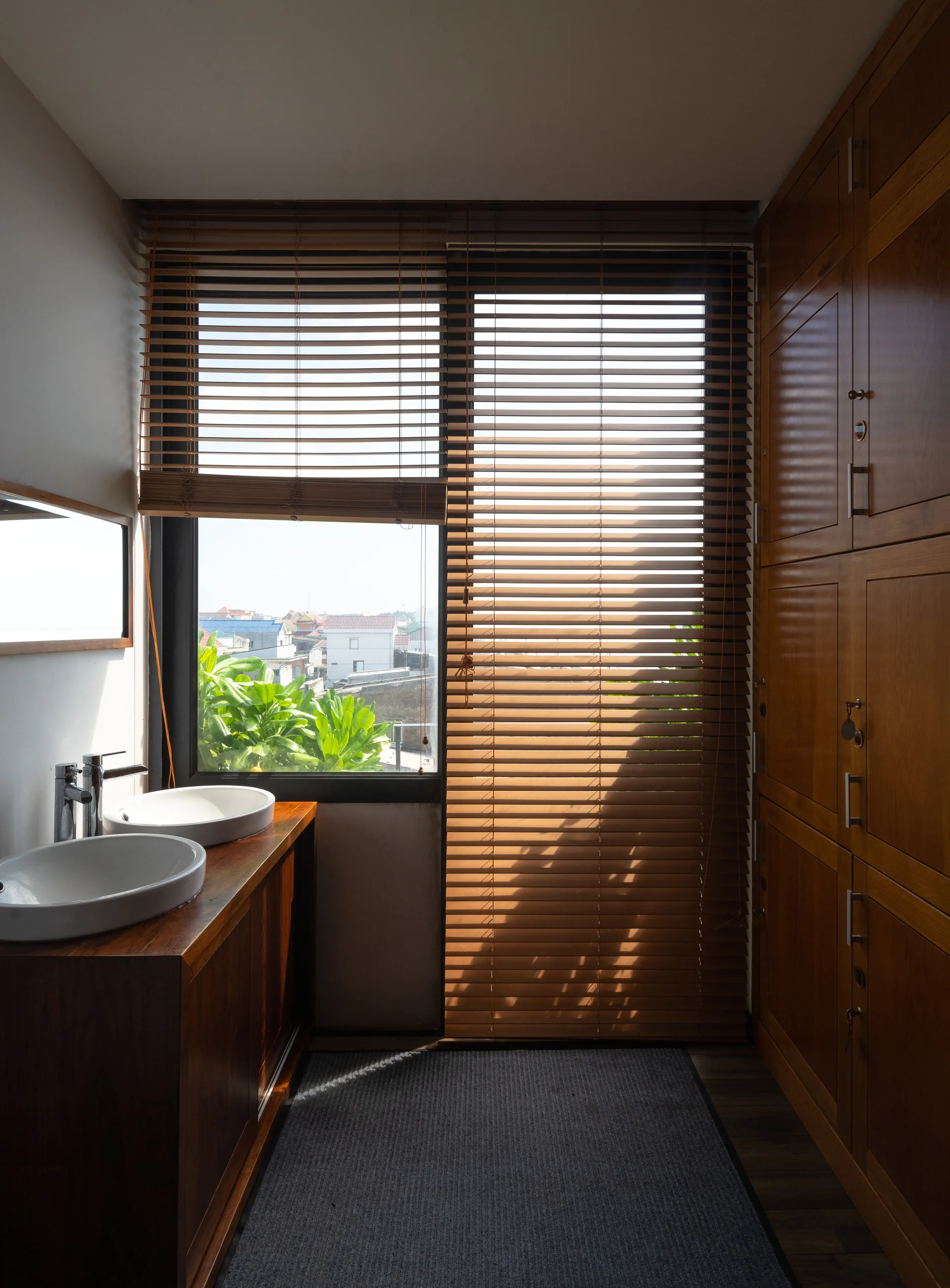 A narrow bathroom features a wooden double-sink vanity, a tall wood storage cabinet, and a large window with horizontal wood blinds.