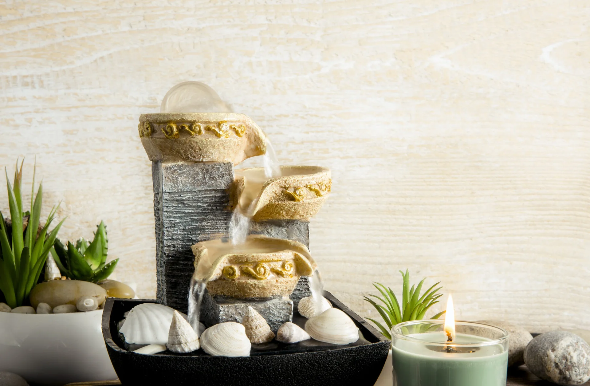 A three-tier tabletop fountain with cascading water sits beside a lit green candle, potted succulents, and scattered seashells against a light wood background.