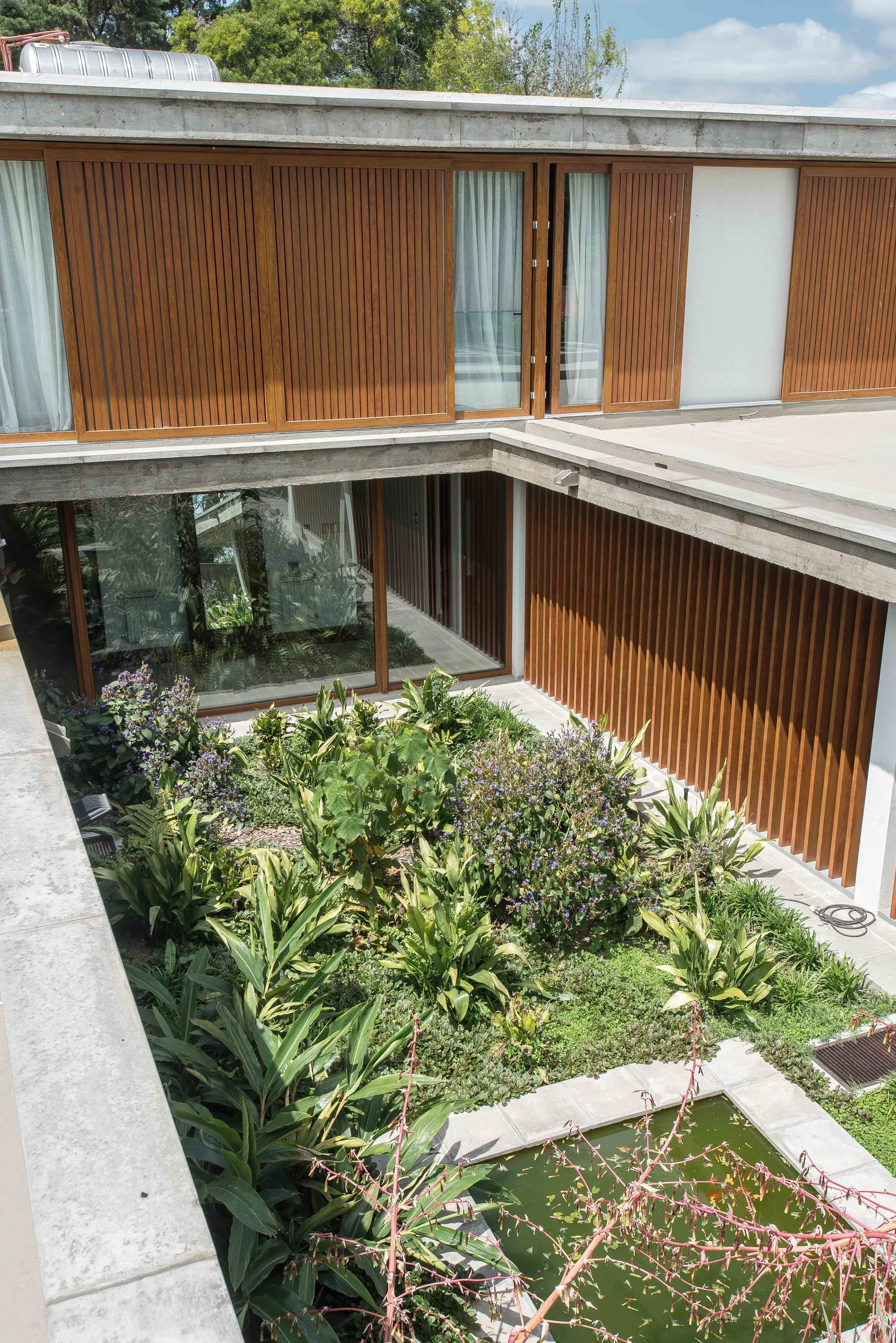 A modern residential courtyard features a lush planted garden with a small pond surrounded by concrete structures and vertical wood slat siding.