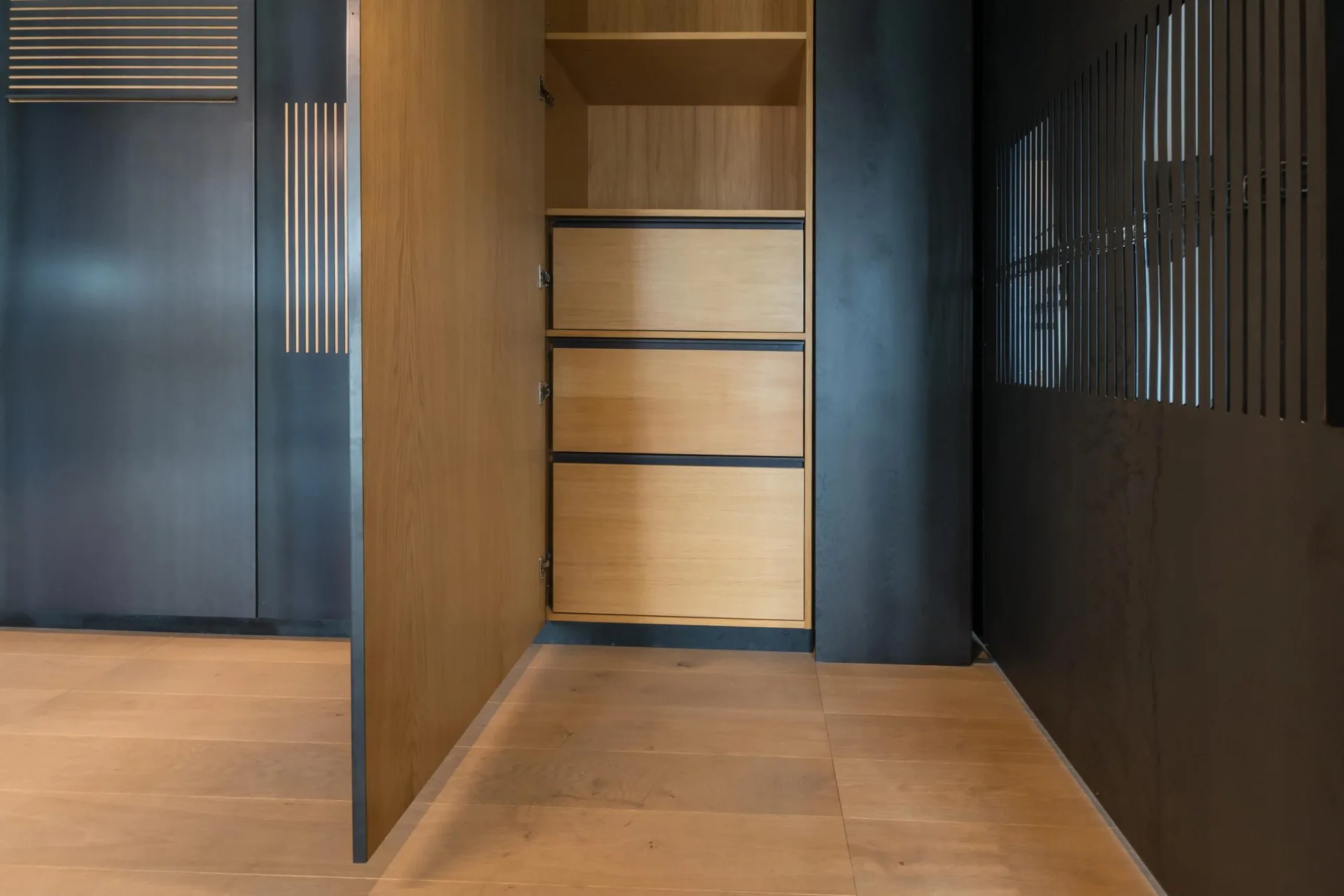 An open cabinet door reveals a natural wood interior with stacked drawers and shelving, contrasted by dark charcoal outer panels and wood flooring.