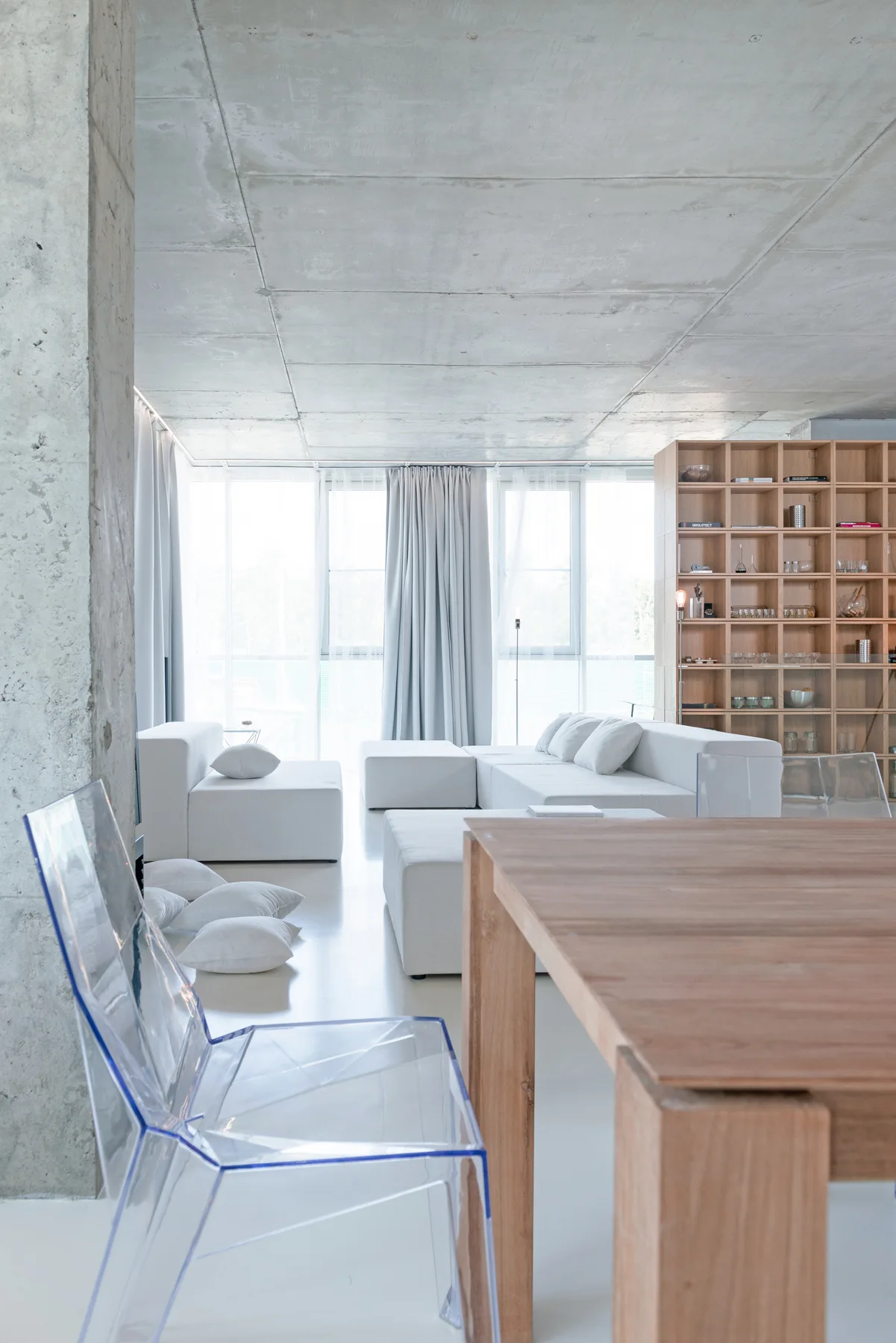 A minimalist room features an exposed concrete ceiling, a white modular sofa, light grey curtains, and a wooden dining table in the foreground.