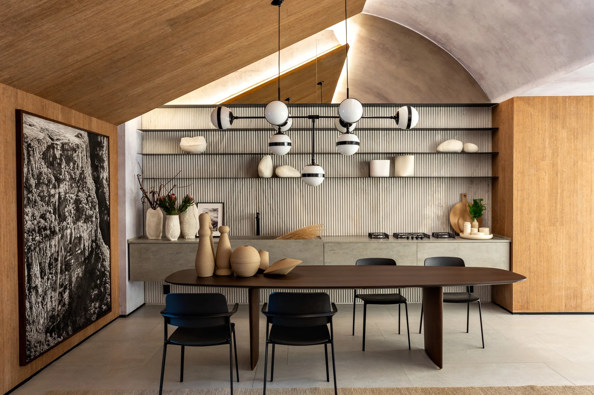 A minimalist dining area featuring a dark wood oval table, black chairs, a fluted accent wall with open shelving, and a vaulted wood ceiling.