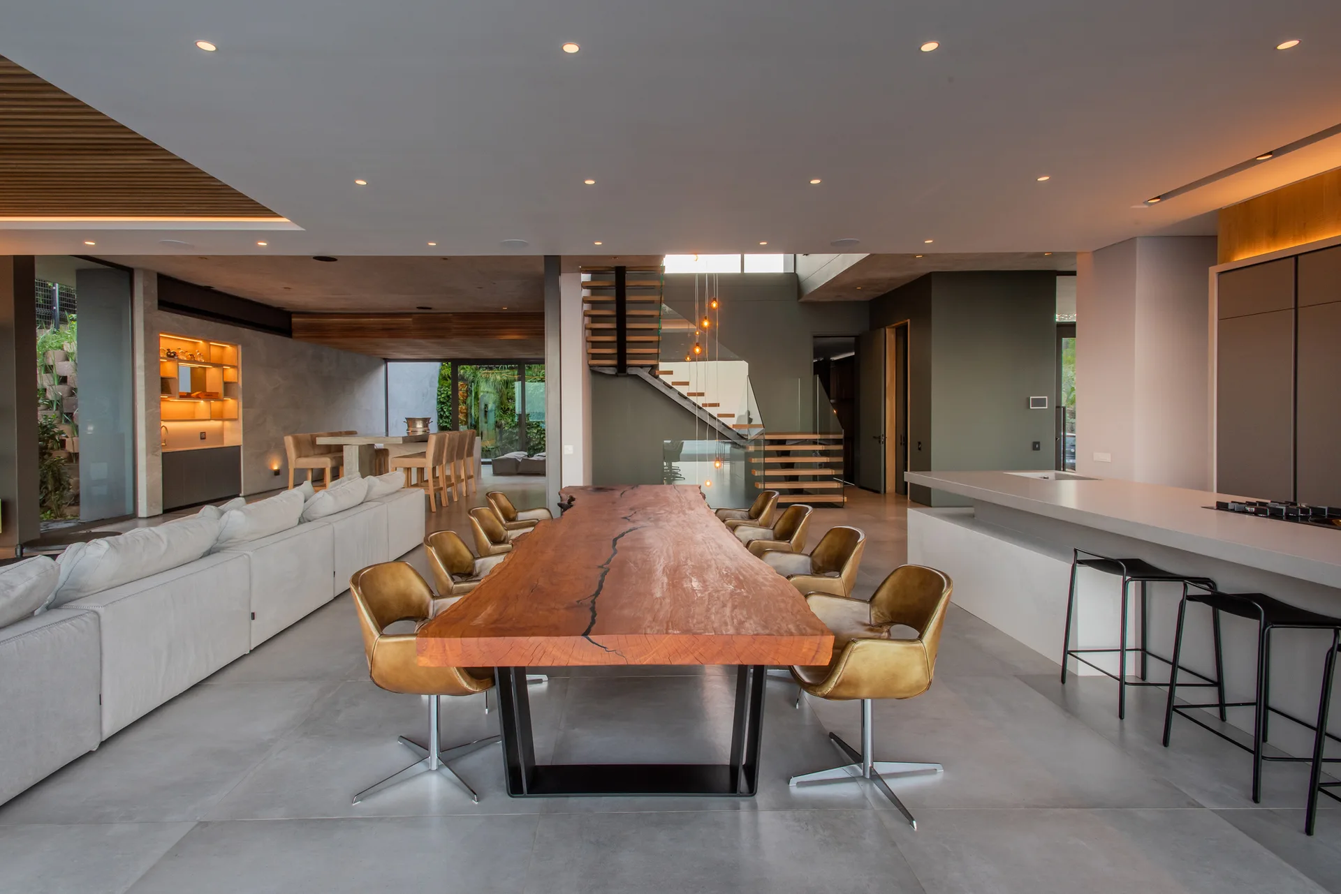 A spacious modern dining room featuring a long live-edge wood table, camel leather swivel chairs, a large kitchen island, and an open riser staircase.