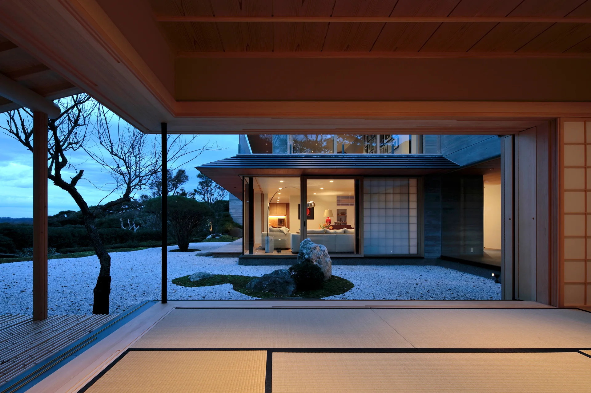A view from a tatami-matted room looking across a gravel courtyard toward a brightly lit living room with sliding glass doors.