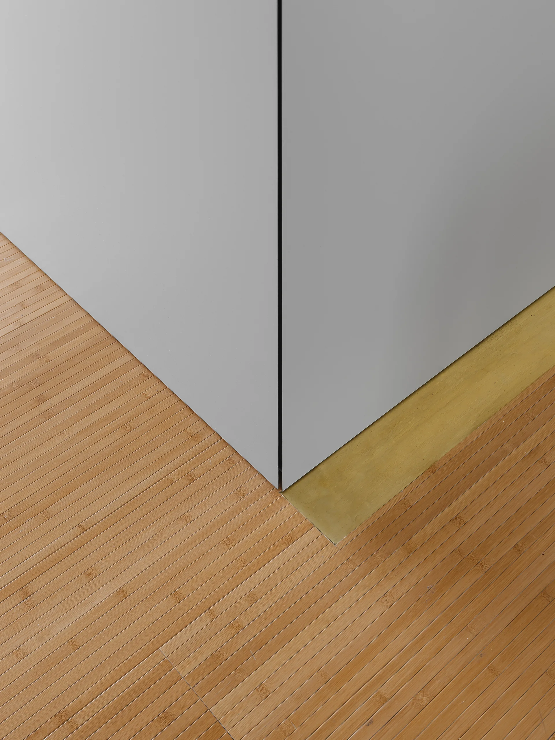 A close-up of a minimalist gray wall corner meeting a slatted bamboo wood floor with an inlaid brass transition strip.