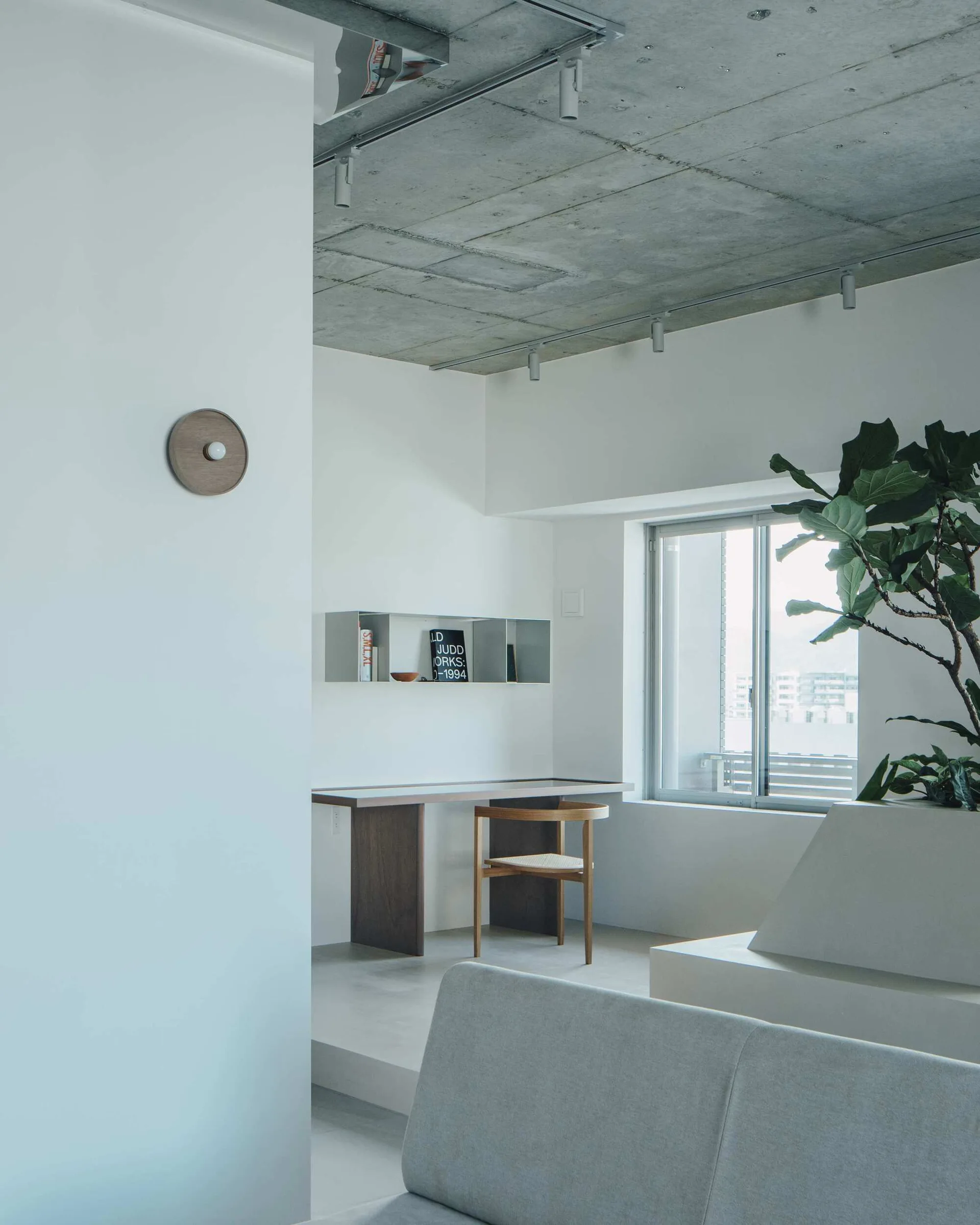 A minimalist living space with an exposed concrete ceiling, a wood writing desk, a cane seat chair, and a large potted plant next to a window.