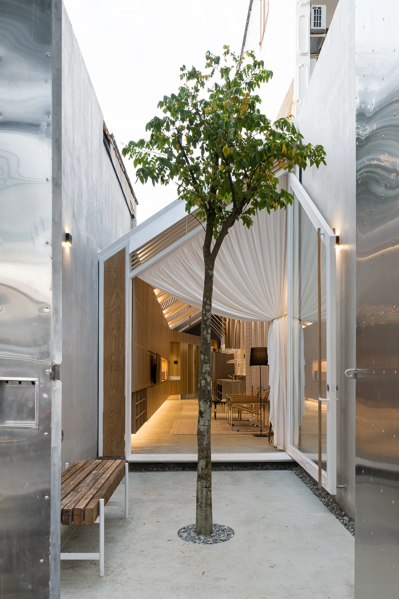 A narrow concrete courtyard features a central tree between metal walls, leading to a modern gabled interior with glass walls and warm wood paneling.