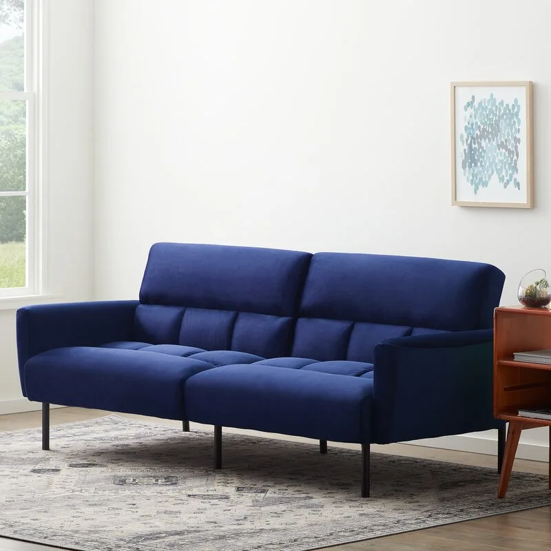 A royal blue tufted sofa with black legs sits on a patterned grey area rug next to a wood side table in front of a white wall.